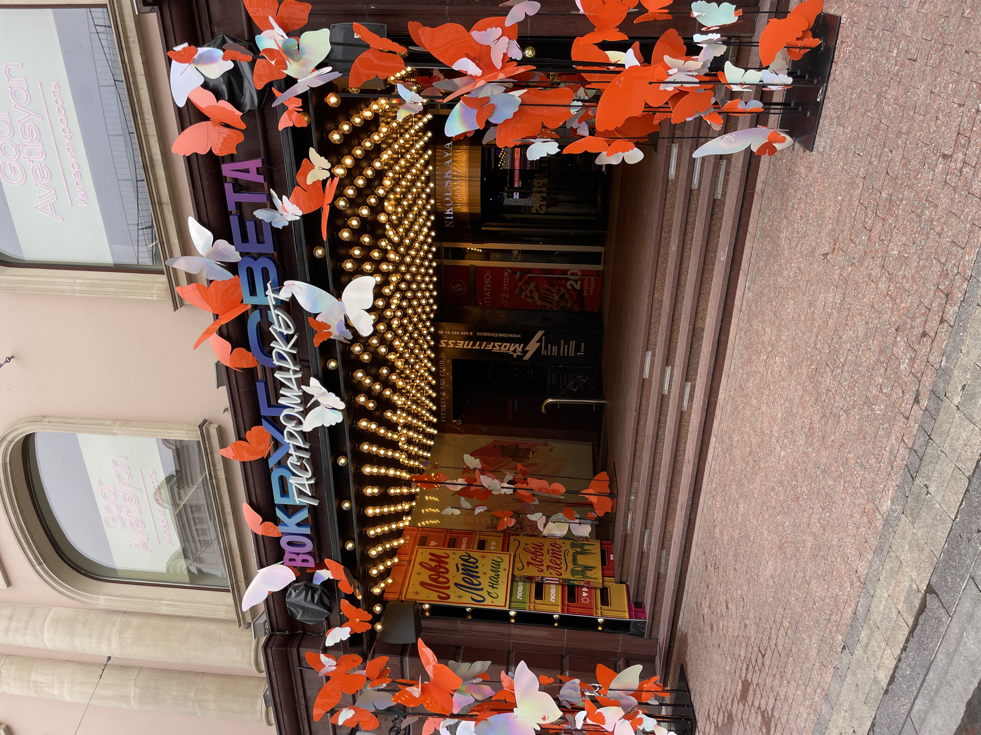 The image depicts the entrance to a building decorated with numerous butterfly-shaped ornaments, predominantly in shades of orange and iridescent white. These ornaments are displayed in clusters and individually, attached to poles and the building's facade. The building has large arched windows above the entrance. One window displays text that appears to read "Goar Avetisyan" and "Империя красоты". The building's signage above the entrance reads "ВОКРУГ СВЕТА". Underneath this, there is a series of lights illuminating the entrance area. A set of stairs leads up to the entrance. To the left of the entrance, there are vertical signs with text, including "Лови Лето". To the right of the entrance, there is a sign that says "NIKOLSKAYA". The ground in front of the building is paved with red and grey stone tiles. The overall scene suggests a festive or decorative display outside a commercial establishment in Moscow, Russia, likely a market or a shopping area.