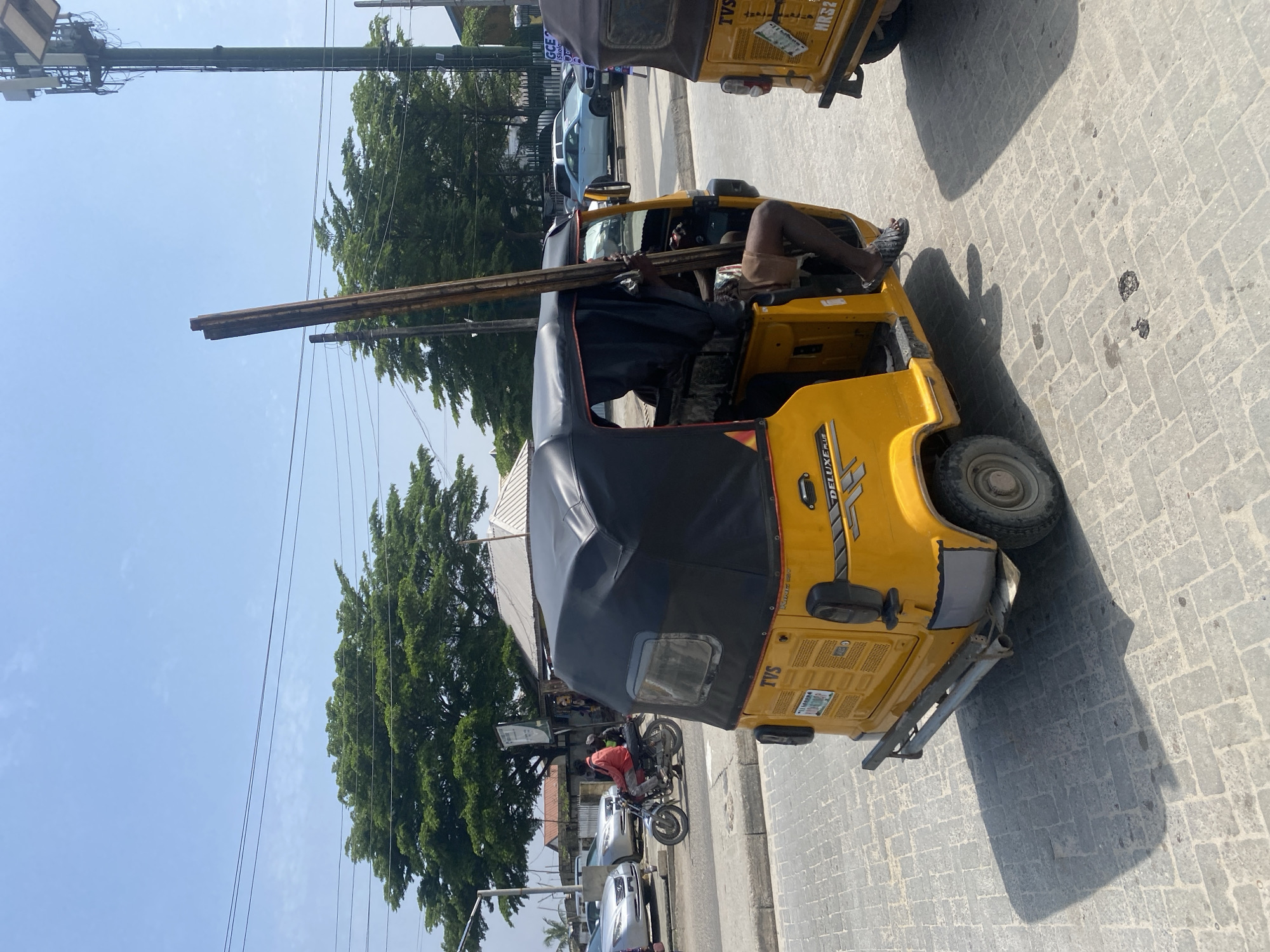 A yellow auto-rickshaw, a "Keke Napep" as commonly known in Nigeria, is parked on a paved street in Ikorodu, Nigeria, under a clear blue sky. The auto-rickshaw, with a black fabric canopy, has the word "DELUXE PLUS" on its side. A person, wearing brown shorts and sandals, is partially visible inside the driver's seat, holding a long, thin, wooden pole. Another auto-rickshaw is parked to the right, partially visible. In the background, there are trees, power lines, a cell phone tower, and several cars on the street. A person on a motorcycle is visible in the distance. The ground is paved with light gray rectangular stones.