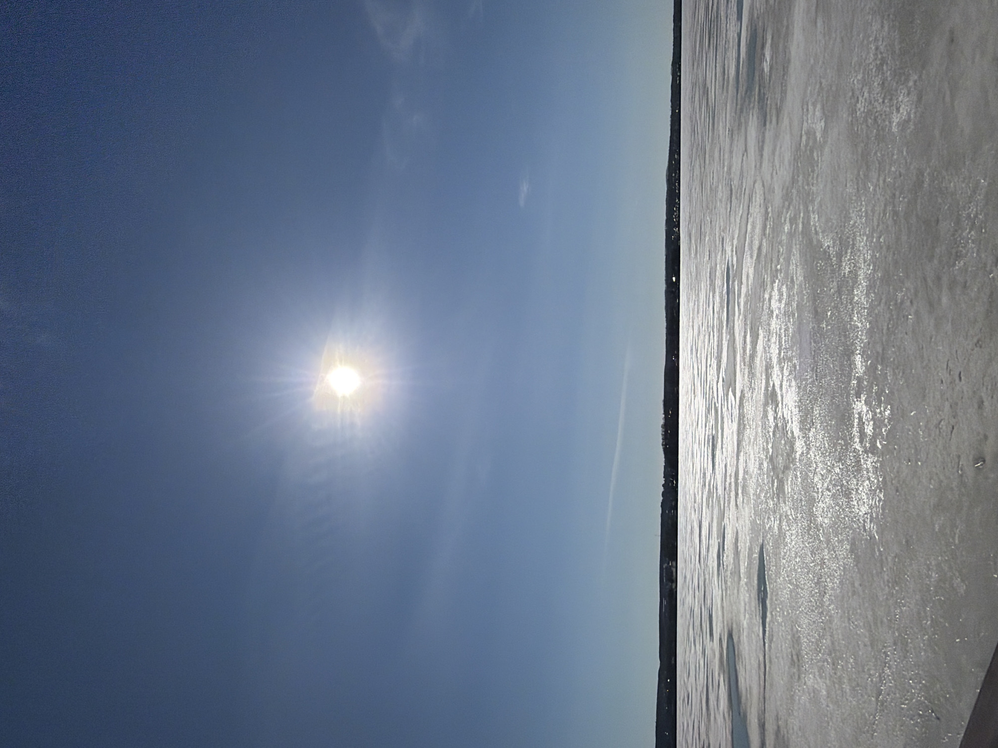 The image depicts an expansive outdoor winter landscape, featuring a bright sky and a large frozen body of water under clear conditions. The horizon line stretches across the middle of the frame, dividing the scene.
The upper portion of the image is dominated by a clear, blue sky, with the sun shining intensely in the upper left. The sun appears as a brilliant, radiant disc, casting bright rays and producing a noticeable lens flare. A few faint, wispy clouds are subtly visible against the blue backdrop.
Below the horizon, the foreground consists of a vast, textured surface that strongly suggests a frozen body of water, such as a lake or reservoir. Given the location context of Votkinsk, Russia, this is likely the frozen Votkinsk Reservoir. This icy surface is highly reflective, with silvery-gray tones highlighting where the sunlight catches its uneven textures. The varying patterns indicate possibly rippled ice, snow, or refrozen slush. A dark, slender line, representing a distant shoreline or land, separates the ice from the sky at the horizon.
No people, distinct objects, or specific activities are visible in the photograph. The bright sunshine indicates it is daytime, and the overall weather appears clear and crisp. The scene conveys a sense of serene vastness and the stark beauty of a winter landscape. There is no visible text within the image.
