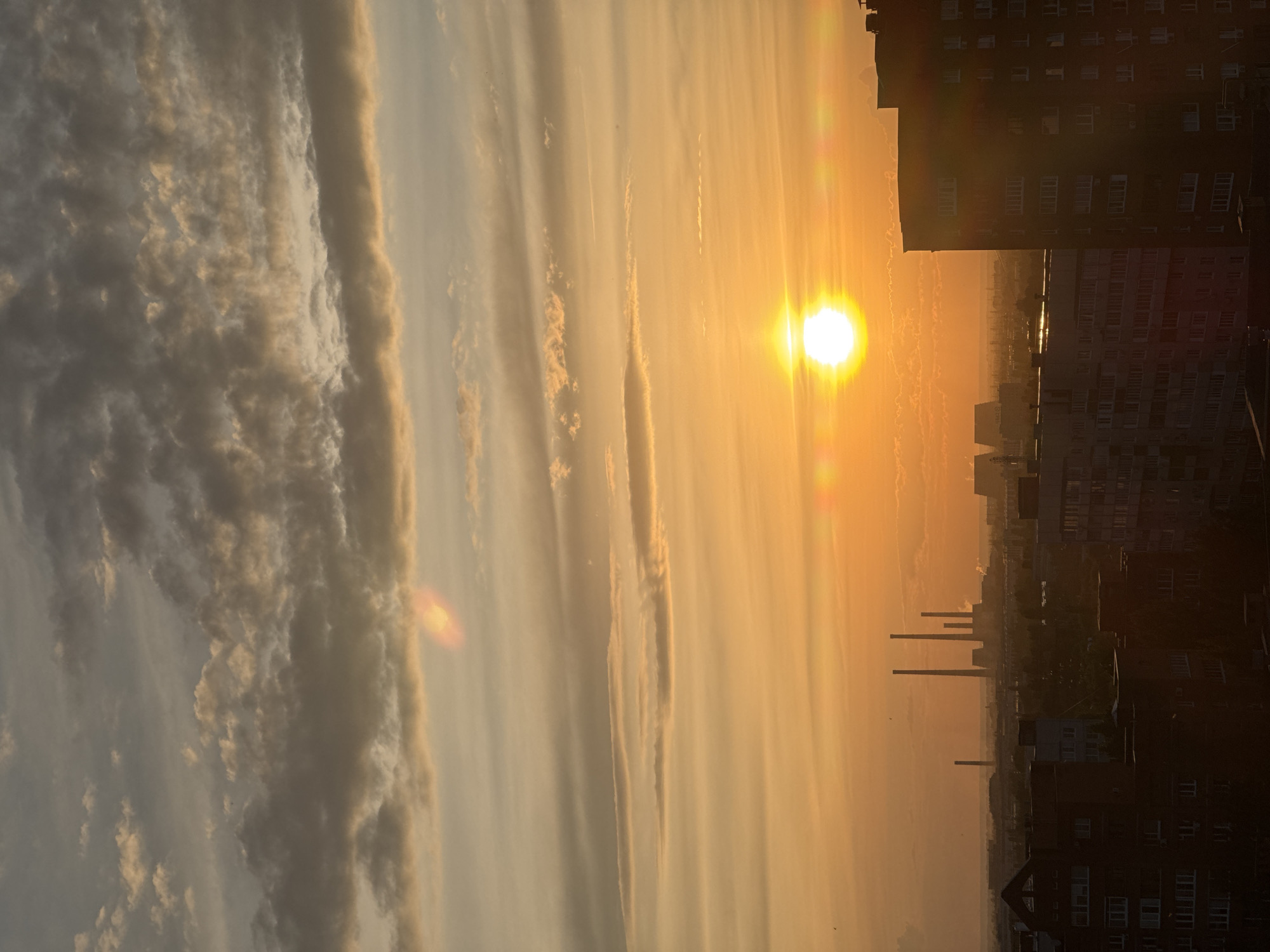 This is a vertical, eye-level shot of a sunset over an industrial cityscape. The sun, a bright white orb, is located slightly left of center in the upper half of the frame, casting a warm, orange glow across the sky. Wispy clouds streak across the sky, some tinged with pink and orange from the setting sun. In the foreground, silhouetted buildings of various heights create a dark, jagged skyline. To the left and slightly further back, a cluster of tall smokestacks, likely from a power plant or factory, pierce the horizon. The overall impression is one of industrial activity juxtaposed with the natural beauty of the sunset.