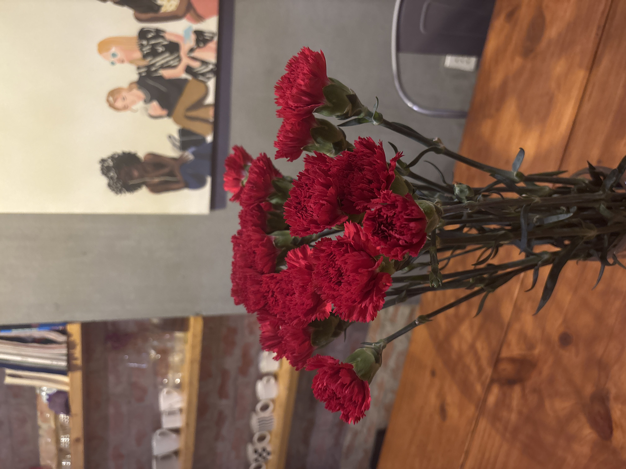 A tilted eye-level shot reveals a bouquet of vibrant red carnations prominently displayed on a wooden table. The flowers' stems extend rightwards across the table. A portion of the background shows a bookshelf with various items. To the left, a framed artwork adorns the wall, featuring painted figures. Further back, a grey wall and the back of a chair can be seen. The location is likely Đà Nẵng, Vietnam, though this detail does not significantly alter the visual information.
