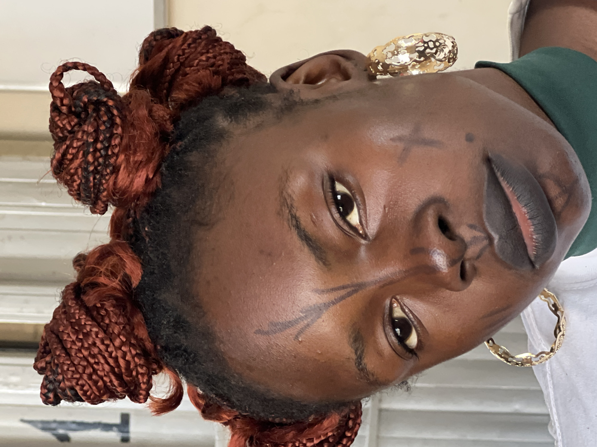 The image is a close-up portrait of a young woman with dark skin. She has her hair styled in numerous braided buns, some appearing reddish-brown. She is wearing large, ornate gold hoop earrings. Her face is adorned with dark markings; there is a V-shaped design on her forehead and a cross-like marking on her cheek. She is wearing dark lipstick. The background consists of vertical white and light-colored elements, with a dark numeral '1' visible on the left. The overall lighting suggests an indoor setting. The specific location in Jos, Nigeria, cannot be determined from the visual information provided in the image.