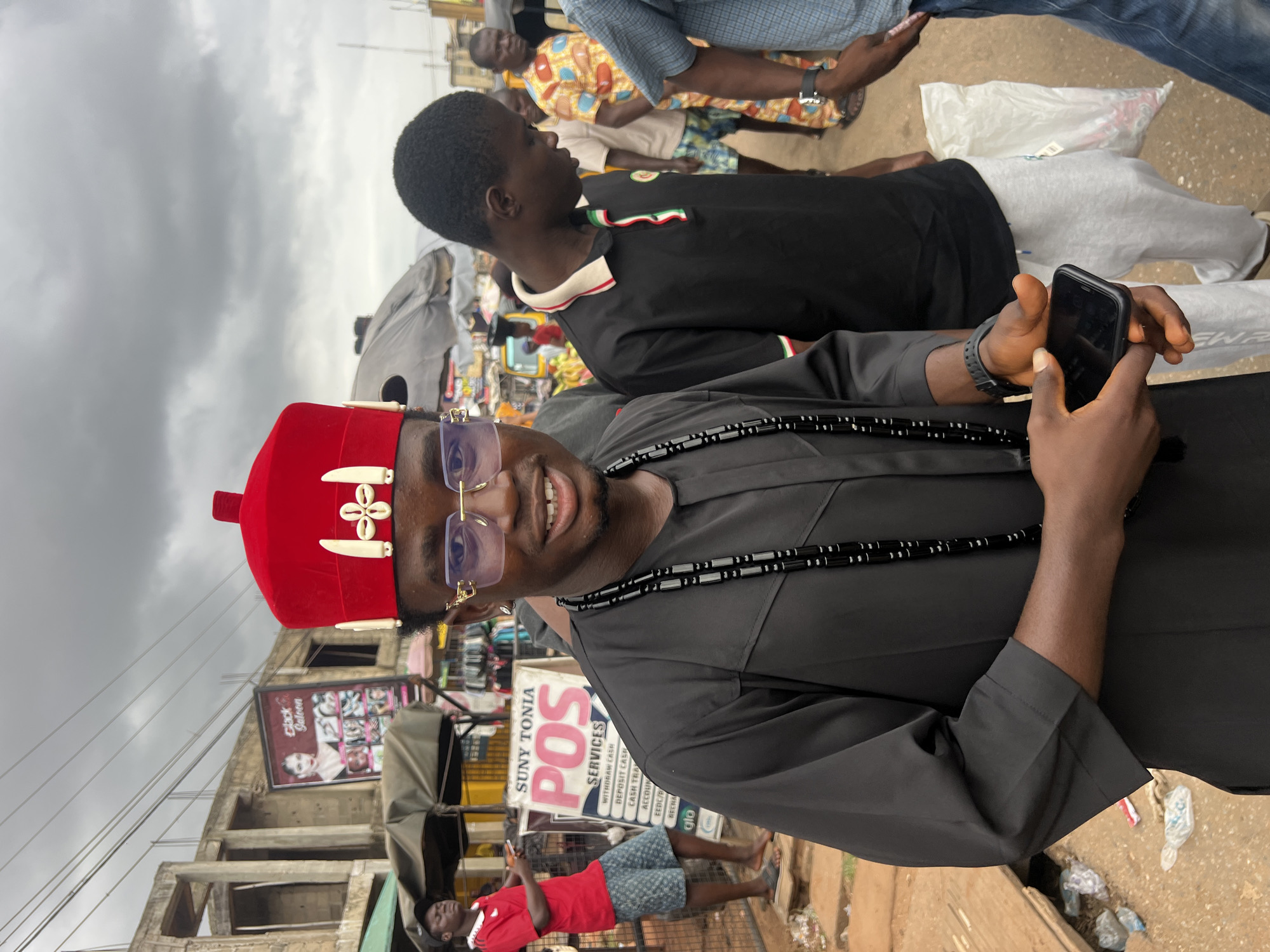 In Nkpor, Nigeria, an outdoor scene unfolds under a cloudy sky. A Black male, center-frame, is dressed in a traditional black garment and a bright red hat adorned with white decorative elements. He wears gold-rimmed glasses and a black beaded necklace. He holds a smartphone in his right hand, his index finger extended as if operating it. To his right, another Black male in a black polo shirt and grey sweatpants looks down. In the background, several other individuals are visible, as are market stalls and buildings. A prominent sign behind the main subject reads "SUNY TONIA POS SERVICES". The overall environment appears to be a busy street or market area.
