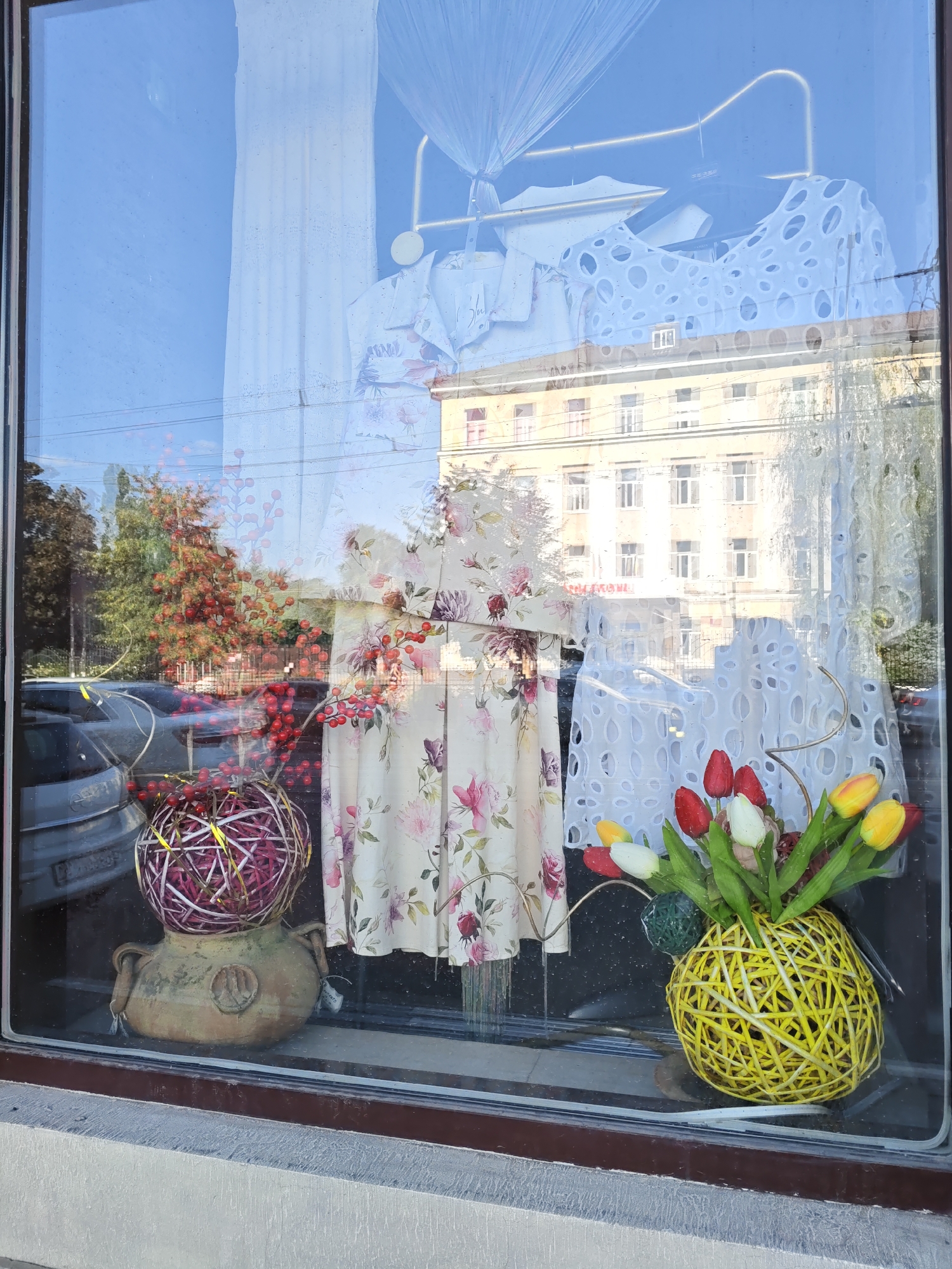 The image shows a storefront window display in Rostov-on-Don, Russia. The window reflects the sky, buildings, and trees. Inside the display, there are several articles of clothing hanging on a rack, including a white lace top with circular cutouts and a long dress with a floral pattern. In front of the clothing, there are two decorative spherical objects. One is a terracotta pot topped with a round, woven object in shades of purple and gold. The other is a yellow woven spherical vase holding a bouquet of tulips in red, white, and yellow. The display is viewed from the exterior of the shop.