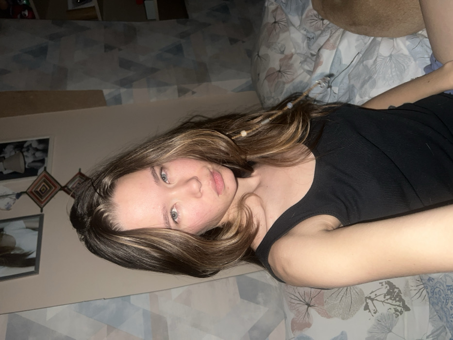 A young woman lies on a bed with a floral patterned blanket. She wears a black tank top. Her light brown hair has blonde highlights and decorative beads woven into it. She has fair skin and blue eyes. The background includes wall decorations, possibly photos or tapestries, and a patterned wall. The image appears to be a casual, possibly self-taken, photograph.