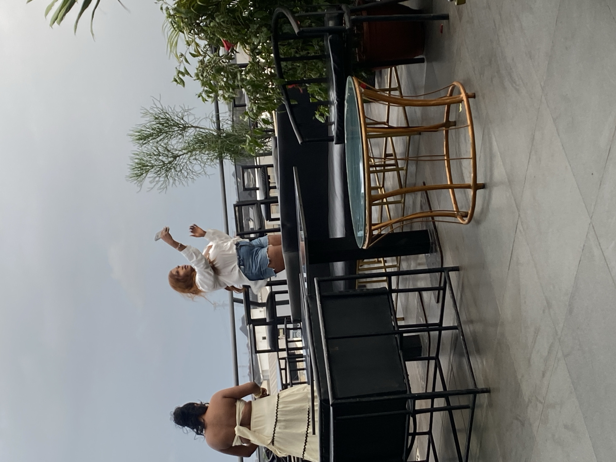 Two women are standing on a rooftop patio. The woman on the left is wearing a strapless cream-colored dress with a ruffled hem and is facing away from the camera. The woman on the right is wearing a white long-sleeved shirt and denim shorts, and she is holding a drink in her right hand while looking up at the sky. The patio is furnished with black metal chairs and tables with black cushions. There is a small round table with a gold base and a glass top in the foreground. Plants are visible in the background, along with a cloudy sky and the tops of buildings.