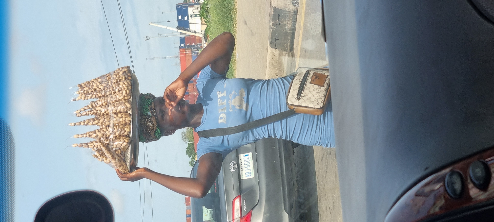 A woman balances a tray of peanuts on her head in Satellite Town, Nigeria. She wears a light blue dress and a patterned headwrap, and a crossbody bag hangs on her side. She is standing next to a gray Toyota Camry with the license plate "ABJ 660 CW." Cargo cranes and shipping containers are visible in the background. The scene is viewed from inside a vehicle, with a portion of the dashboard visible.