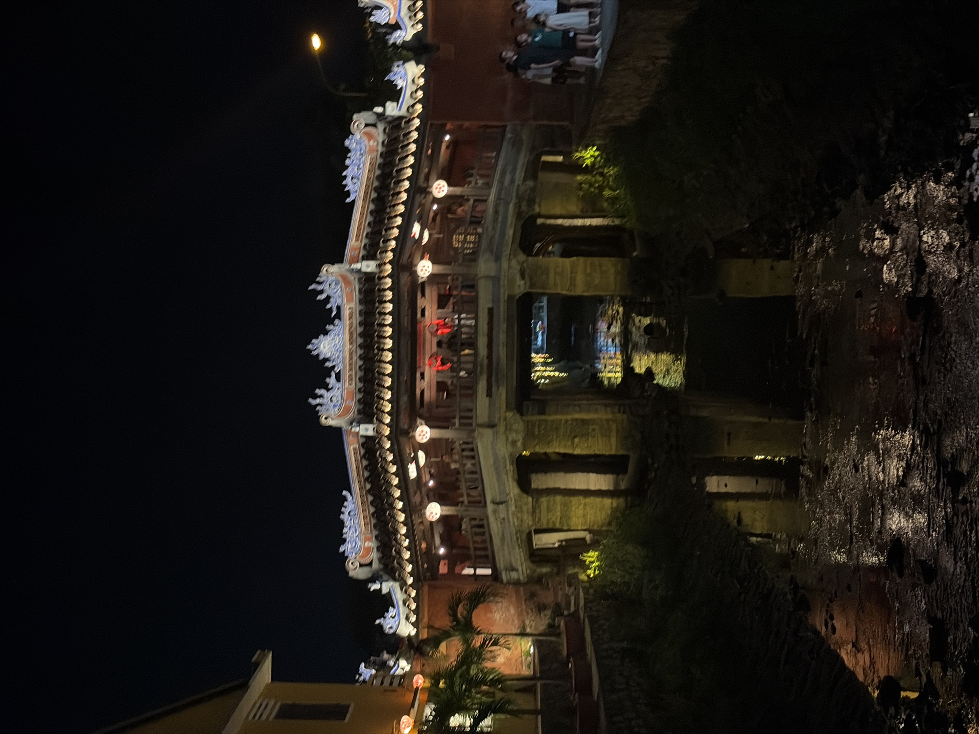 The image displays a captivating nighttime view of the Japanese Covered Bridge (Chùa Cầu) in Hội An, Vietnam. The prominent structure spans a narrow waterway, its traditional ornate roofline and intricate carvings illuminated by warm-toned lights and spherical lanterns that hang along its length. These lights cast soft reflections onto the dark water below, creating a tranquil atmosphere.
The time of day is clearly night, with the sky appearing completely dark and the scene relying entirely on artificial illumination. A single street light glows in the upper left portion of the frame, further contributing to the artificial lighting of the area.
Several people are visible in the scene. A small group of individuals stands on the pathway or bank to the right of the bridge, and more figures can be discerned within the illuminated sections of the bridge itself, suggesting an active, though not crowded, evening at this popular landmark. Their activity appears to be casual sightseeing or strolling.
The surrounding environment includes dark foliage and trees along the banks, dimly lit by the ambient light, and hints of other buildings, such as a yellowish structure on the far left. The overall impression is one of serene beauty, highlighting the bridge as a focal point. There is no discernible text visible in the image.