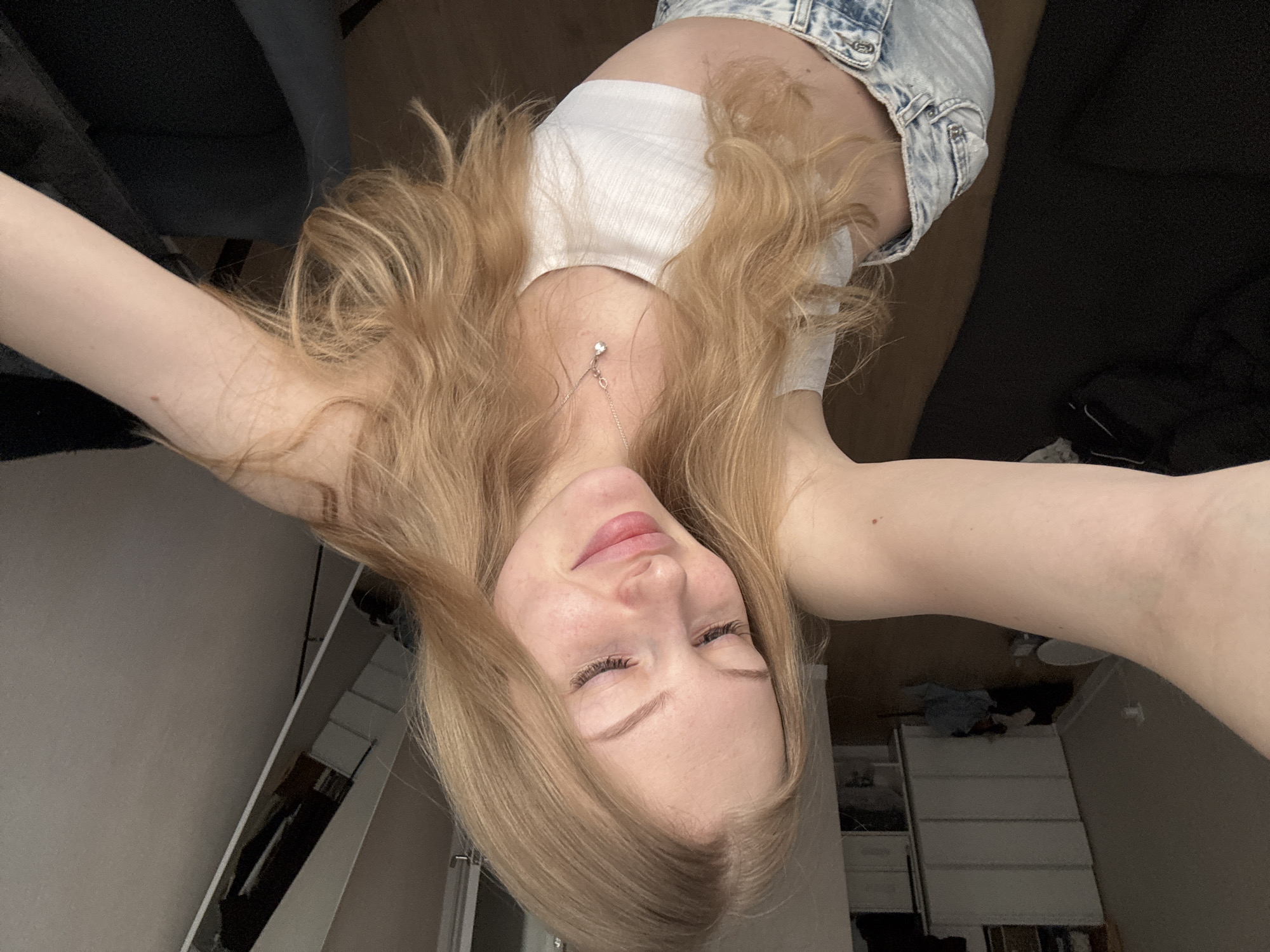 A young woman with long blonde hair is taking a selfie from a high angle. She is wearing a white crop top and denim shorts. She is indoors, likely in a bedroom, as evidenced by the dresser in the background and the furnishings. The location in the city of Perm, Russia is not evident from this image.