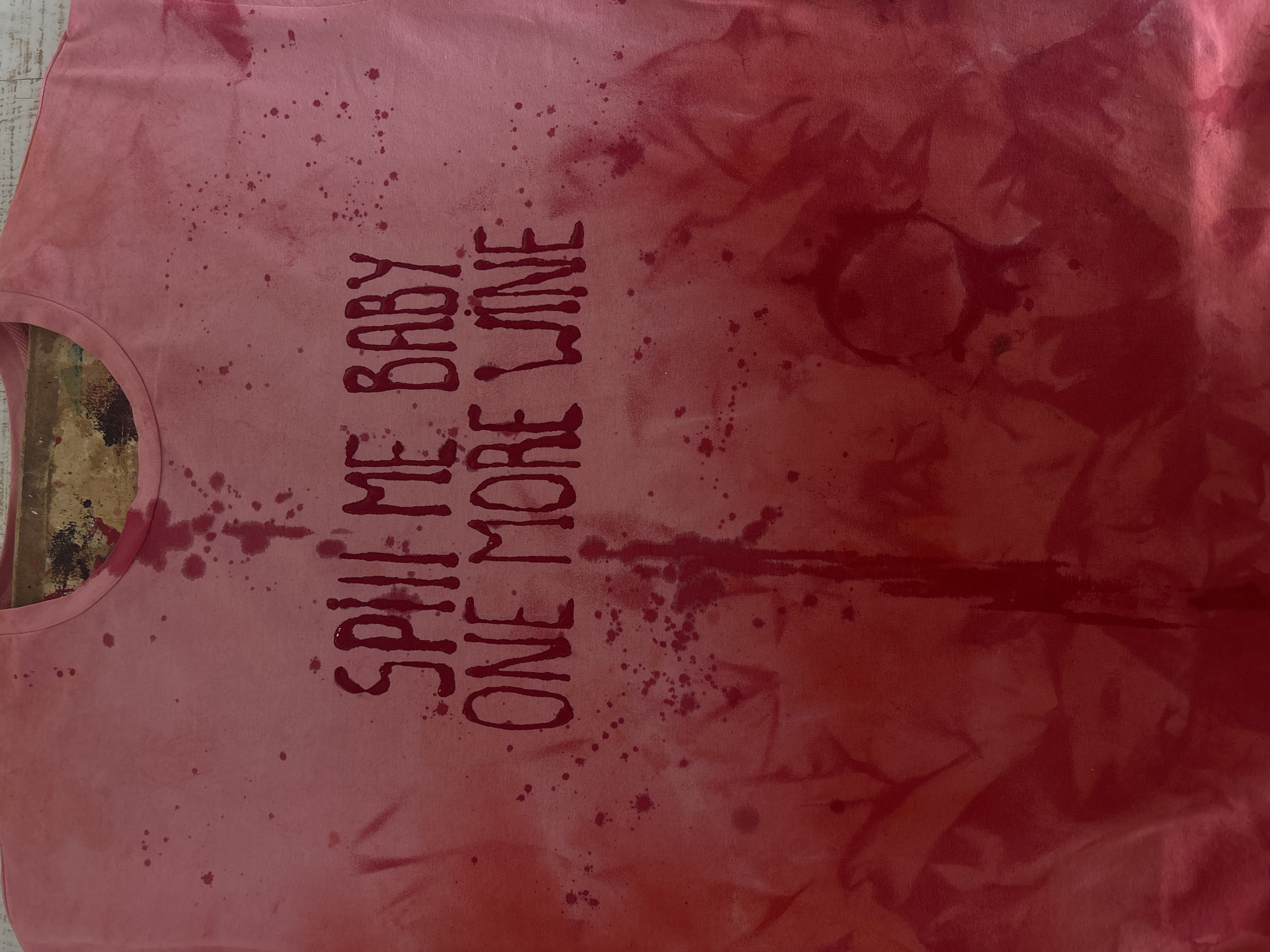 A pink t-shirt is lying flat on a surface. The phrase "SPILL ME BABY ONE MORE WINE" is written vertically in a darker red hue across the front. Splatters of the darker red hue are scattered across the t-shirt, with larger concentrations and drips towards the right side of the shirt. The t-shirt is near a window where wood flooring is visible.
The location is not discernible from the image. There is no evidence to suggest this picture was taken in Krasnoyarsk, Russia.