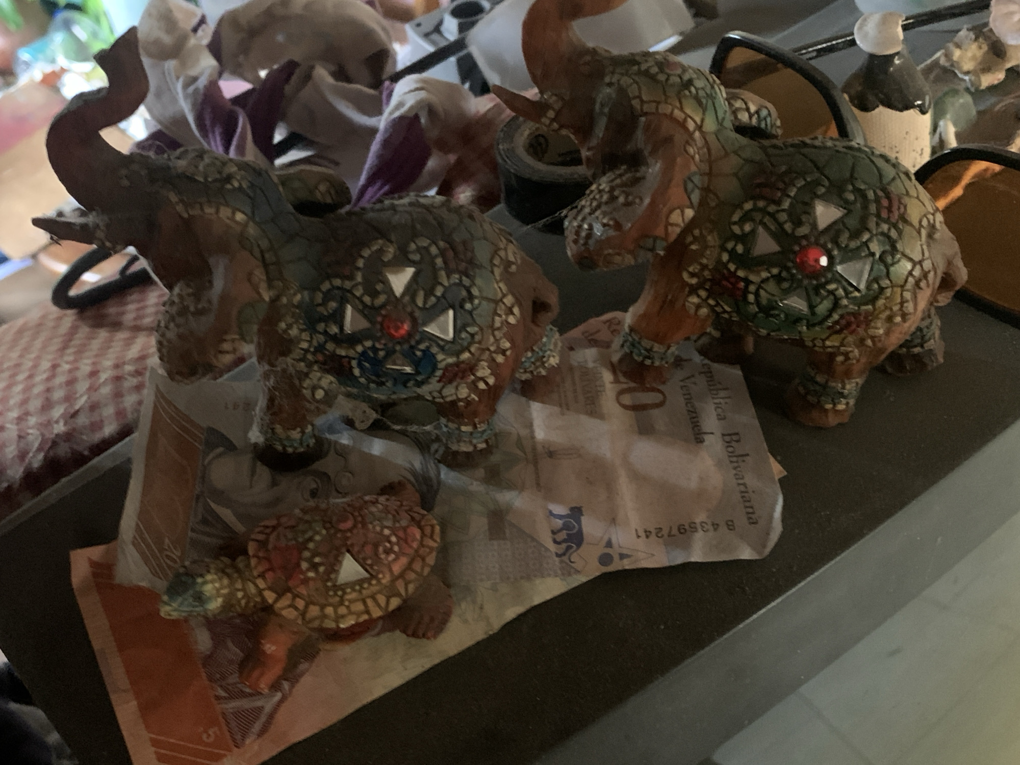The image shows two decorative elephant figurines and a turtle figurine sitting on top of a Venezuelan Bolívar banknote. The elephants are decorated with colorful patterns and have raised trunks. The turtle has a reddish-brown shell with geometric designs. The banknote shows the words "República Bolivariana de Venezuela". The location in Santa Ana, Venezuela is not discernible.