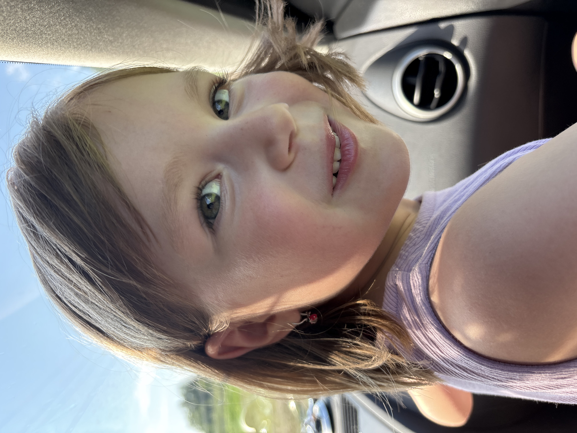 The image is a close-up portrait of a young girl with brown hair and green eyes, seen from the chest up, in what appears to be the interior of a car. She is wearing a purple tank top and a small dangling earring in her left ear. Sunlight illuminates her face from the side, casting a warm glow. The background is blurred, but hints of greenery and a bright sky suggest an outdoor environment. A car's air vent is visible on the right side of the frame. The girl is looking towards the right of the camera, with a slight smile. The scene is devoid of any context that would place it in the city of Маркидоновка, Russia; no landmarks or identifying features of that specific location are present.