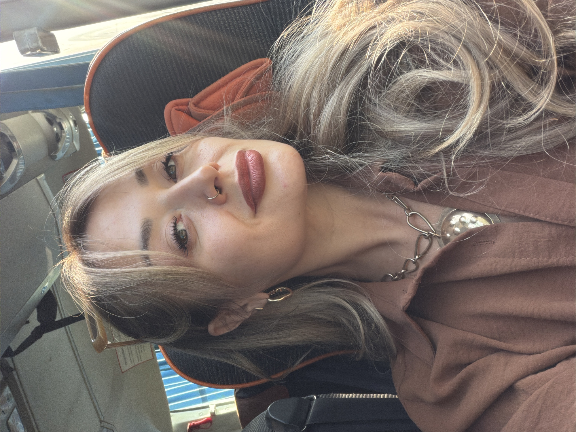 A woman with blonde hair and bangs, wearing a brown shirt and a heart-shaped necklace, is photographed inside a vehicle. She has a nose ring and hoop earrings. The woman is looking towards the camera with a slight smile. The background shows the interior of a bus, with seats and windows visible. The image is taken during the day, with sunlight streaming in, creating a warm glow. The location is the city of Ashkaul, Russia.