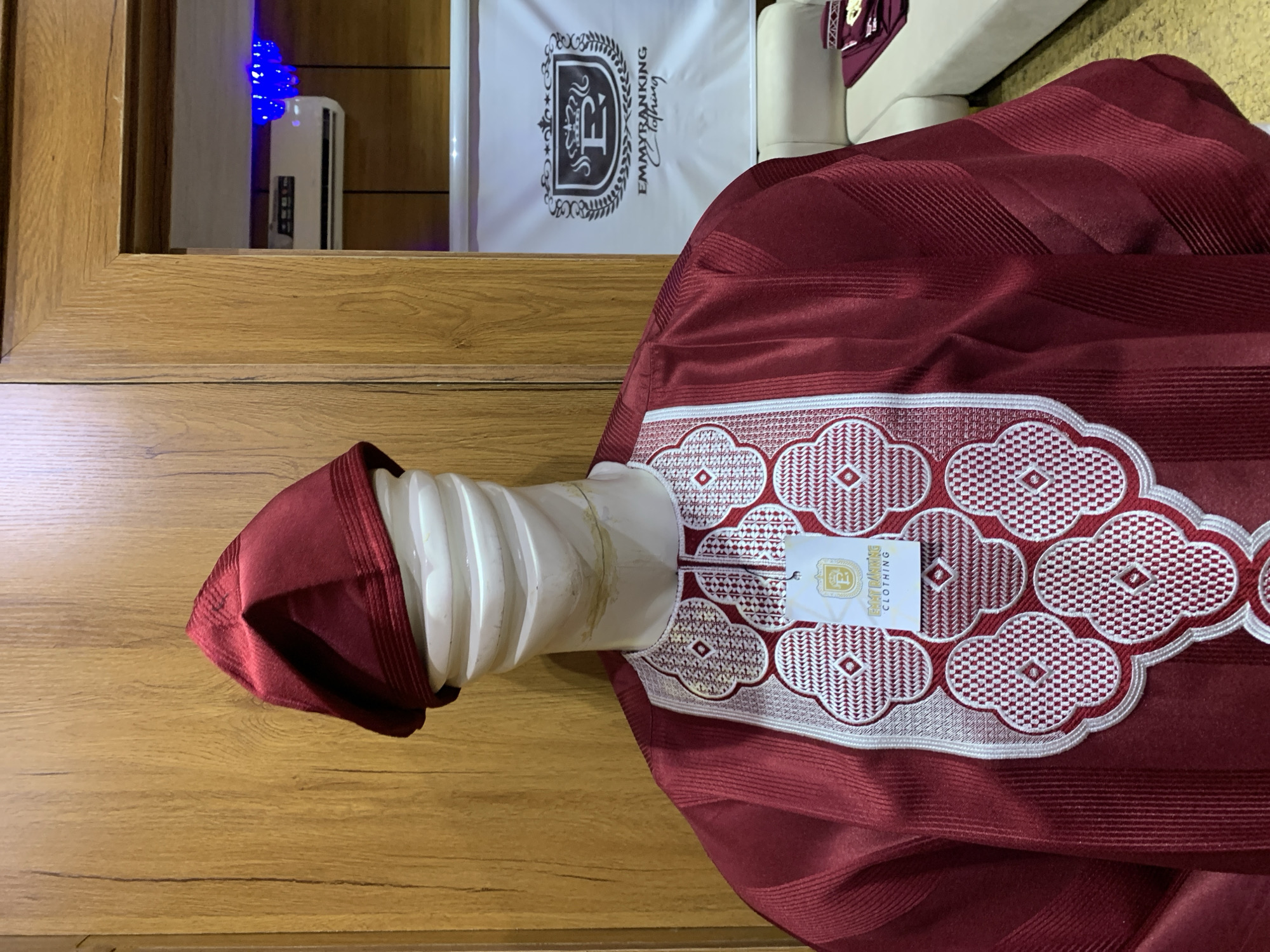 A mannequin is displayed in front of a wooden cabinet in a store. The mannequin wears a maroon traditional Nigerian attire with intricate white and silver embroidery on the chest and a matching maroon cap. A tag with the brand "EMMYRANKING CLOTHING" is attached to the garment. In the background, a white banner with a logo and text is visible, along with a white sofa and what appears to be an air conditioning unit mounted on the wall. The setting suggests a retail environment, possibly a clothing boutique specializing in traditional wear. The location is identified as Gura Top, Nigeria.
