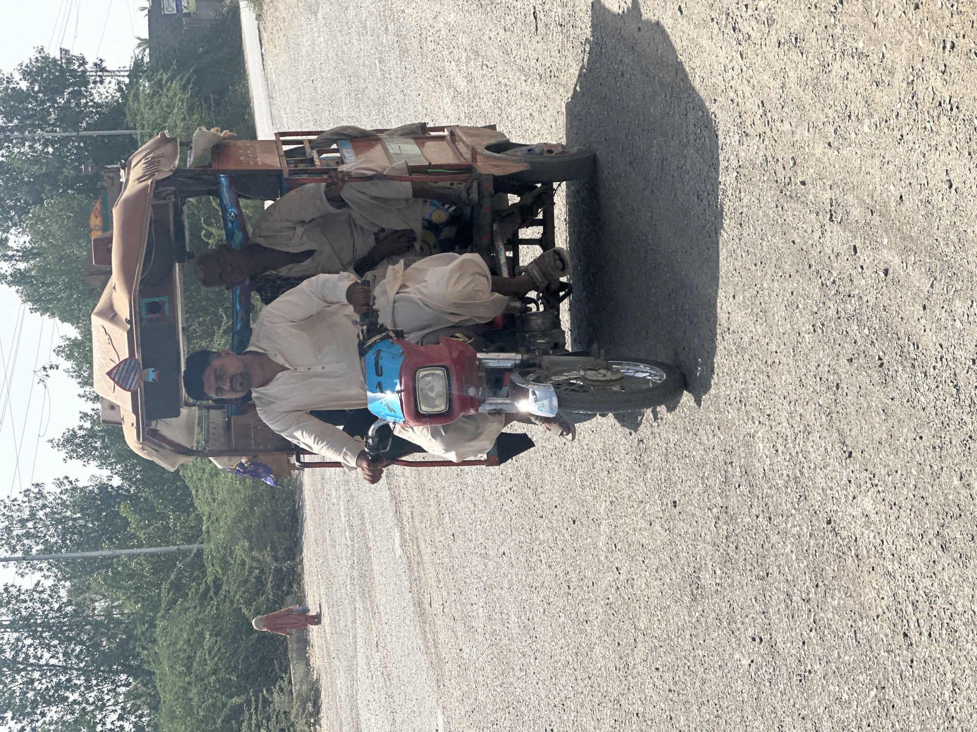 A motorized rickshaw, resembling a motorcycle with an attached passenger carriage, travels along a sunny road. The vehicle's front is red and blue, and it appears to be a modified motorcycle, possibly a Honda 70 or similar model, commonly found in South Asia.
Seated on the motorcycle portion, and acting as the driver, is a man wearing a light-colored shalwar kameez. He is looking directly at the camera with a slight smile. Behind him, several other men, also in traditional light-colored attire, are seated within the open-sided passenger carriage. The carriage itself appears to be made of metal and wood, showing signs of wear and tear, and features some decorative elements.
The setting is a rural or semi-urban environment. The road surface is asphalt or gravel, and a distinct shadow, likely cast by the vehicle or an unseen object, stretches across the road from the right. On the left side of the road, abundant green trees and bushes line the pathway, with a light sky visible above. In the distance on the left, a small figure, possibly a child or woman in a reddish garment, walks on the side of the road. Further back, a building or structure is faintly visible amidst the greenery. The time of day appears to be bright and sunny, indicated by the clear lighting and shadows. No discernible text is visible in the image.