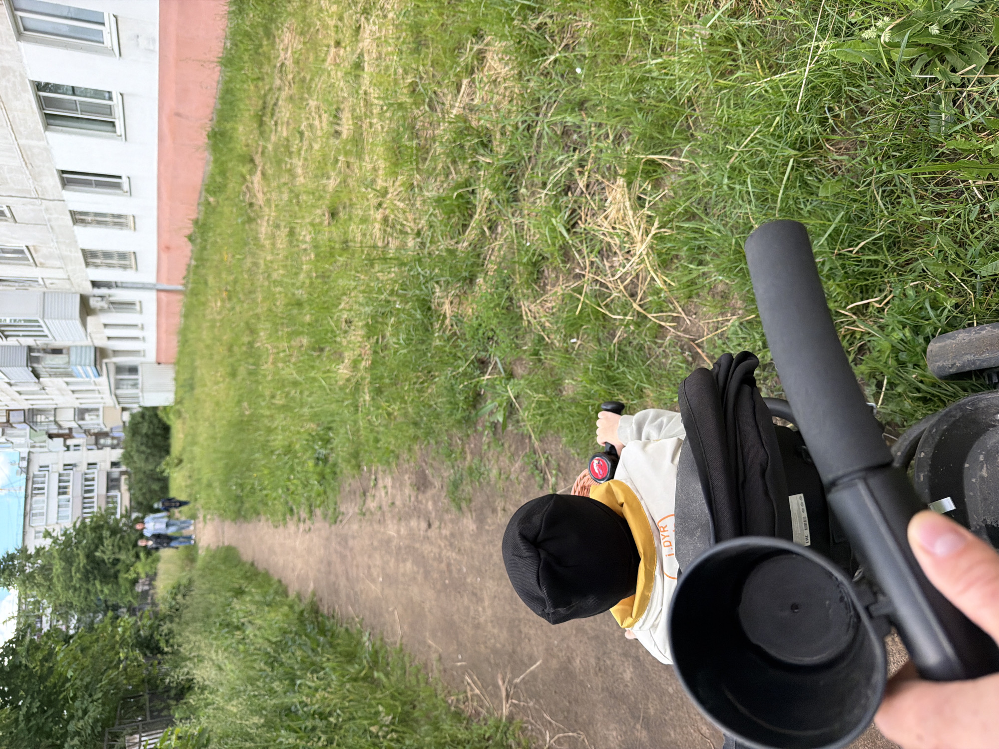 The image, taken from a first-person perspective, captures a scene outdoors on a dirt path next to a grassy area. In the immediate foreground, a hand holds the black, padded handle of what appears to be a stroller or child's tricycle, which also has an attached black cup holder visible. A child is seated in the stroller/tricycle, seen from behind, wearing a black beanie and a light-colored hooded jacket with a yellow lining. Part of the text "i.DYR)" is visible on the back of the child's jacket.
The dirt path stretches into the distance, with green grass and some dry patches flanking it on both sides. On the left side of the path, several multi-story residential buildings, likely apartment blocks, are visible. Further along the path in the mid-ground, a few blurry figures, possibly adults and another child, can be seen walking away from the camera. Trees and other foliage are present further down the path and along the left side.
The scene depicts a casual daytime activity, possibly a walk or outing, in an urban or suburban setting like Chaikovsky, Russia. The lighting suggests an overcast or partly cloudy day, as there are no harsh shadows.