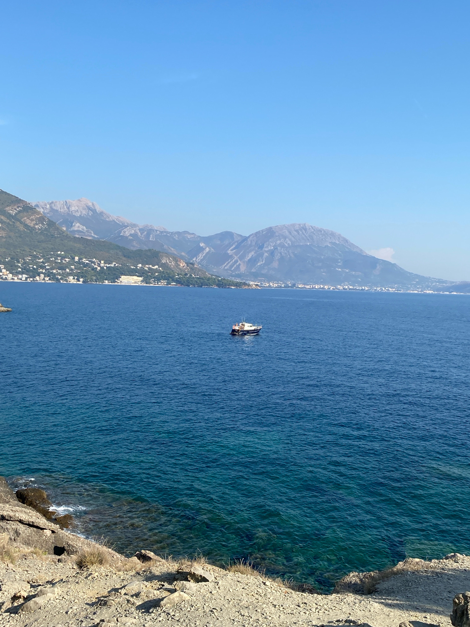 A vertical, eye-level, full shot captures a bright, clear day in Sutomore, Montenegro, overlooking the Adriatic Sea. The foreground is dominated by a rocky, sun-drenched embankment covered with sparse, dry grasses and shadows. The midground is occupied by the deep blue, rippling waters of the sea, with a small, white and dark-hulled boat carrying several passengers visible in the center. The background features a sprawling coastal town with white buildings nestled amongst lush green hills, which rise to meet jagged, grey mountain peaks under a vast, pale blue sky. The scene is peaceful and showcases the natural beauty of the coastal landscape.