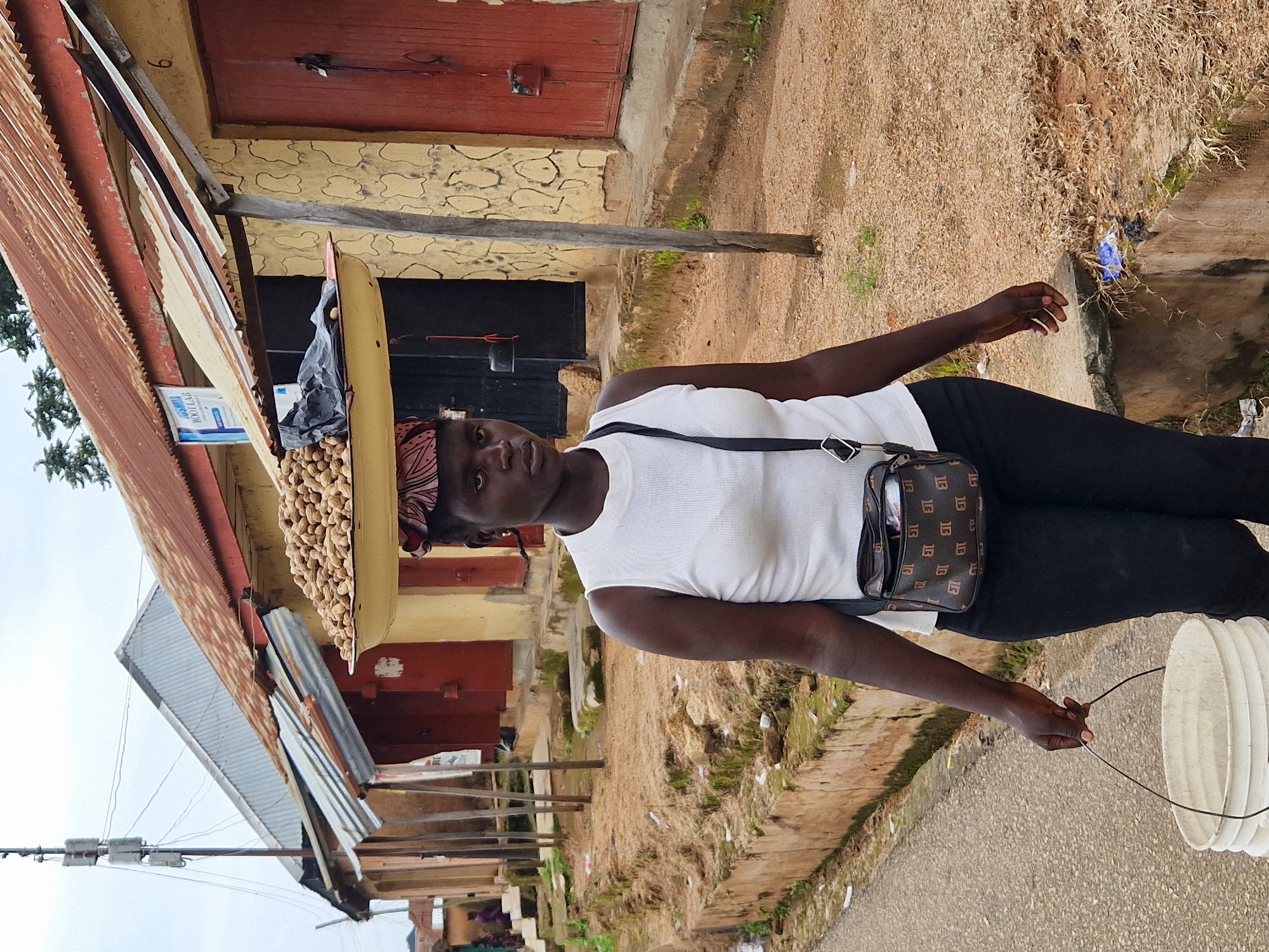A woman in Kabong, Nigeria, walks along a dirt road in front of a row of stalls with corrugated tin roofs. She is wearing a white sleeveless top, black pants, and a patterned headwrap. A tray of peanuts is balanced on her head, and a black bag with a patterned strap is slung across her chest. She is carrying a white bucket by its handle in her left hand. The buildings behind her have yellow and red facades, and some have metal bars on the doors. The ground is covered in dry grass and scattered debris. The sky is overcast.