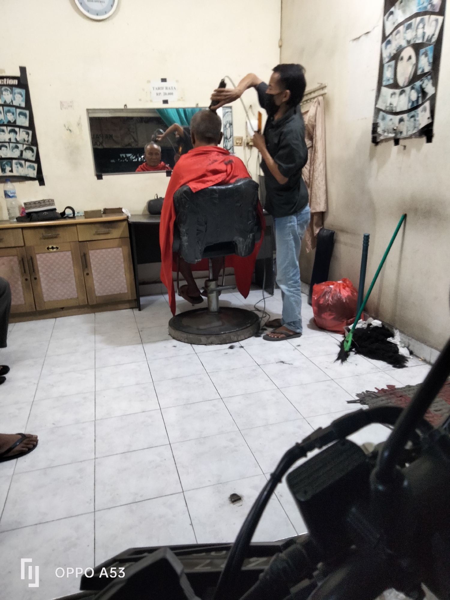 This is a medium-wide shot of a barbershop in Denpasar, Indonesia. A barber, wearing a black shirt, blue jeans, and a black face mask, is using an electric razor to cut the hair of a client seated in a black barber chair. The client is wearing a red cape and a black shirt. The barber shop has white tiled floors and cream-colored walls. On the wall to the left, there is a display of black and white photos of hairstyles, and a sign with the word "ECTION". Above the client, a mirror reflects the barber and client, as well as another person sitting further back in the shop. To the right of the barber, there is a red trash bag, a broom with a green handle, and a pile of black hair on the floor. In the foreground, a portion of a motorcycle is visible, suggesting the photo was taken by someone on a motorcycle. A framed sign with the text "TARIF RATA RP. 20.000" is also visible on the wall.