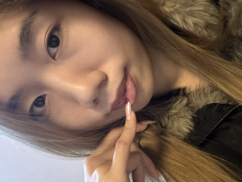 The image depicts a close-up, chest-up view of a young East Asian female, located in Guandu, China. The individual has light brown hair, visible on either side of her face and draping over her shoulders. She wears a dark jacket featuring a prominent grey-brown fur-lined collar. Her left hand is raised, with the index finger touching or very near her lower lip. Her fingernails are long, almond-shaped, and appear to be artificial extensions with a light, semi-transparent base and subtle glitter or speckles. Her lips are slightly parted and appear to have a glossy finish. Her gaze is directed straight towards the viewer. The background is an indistinct, uniformly light-colored surface, suggesting an indoor setting.