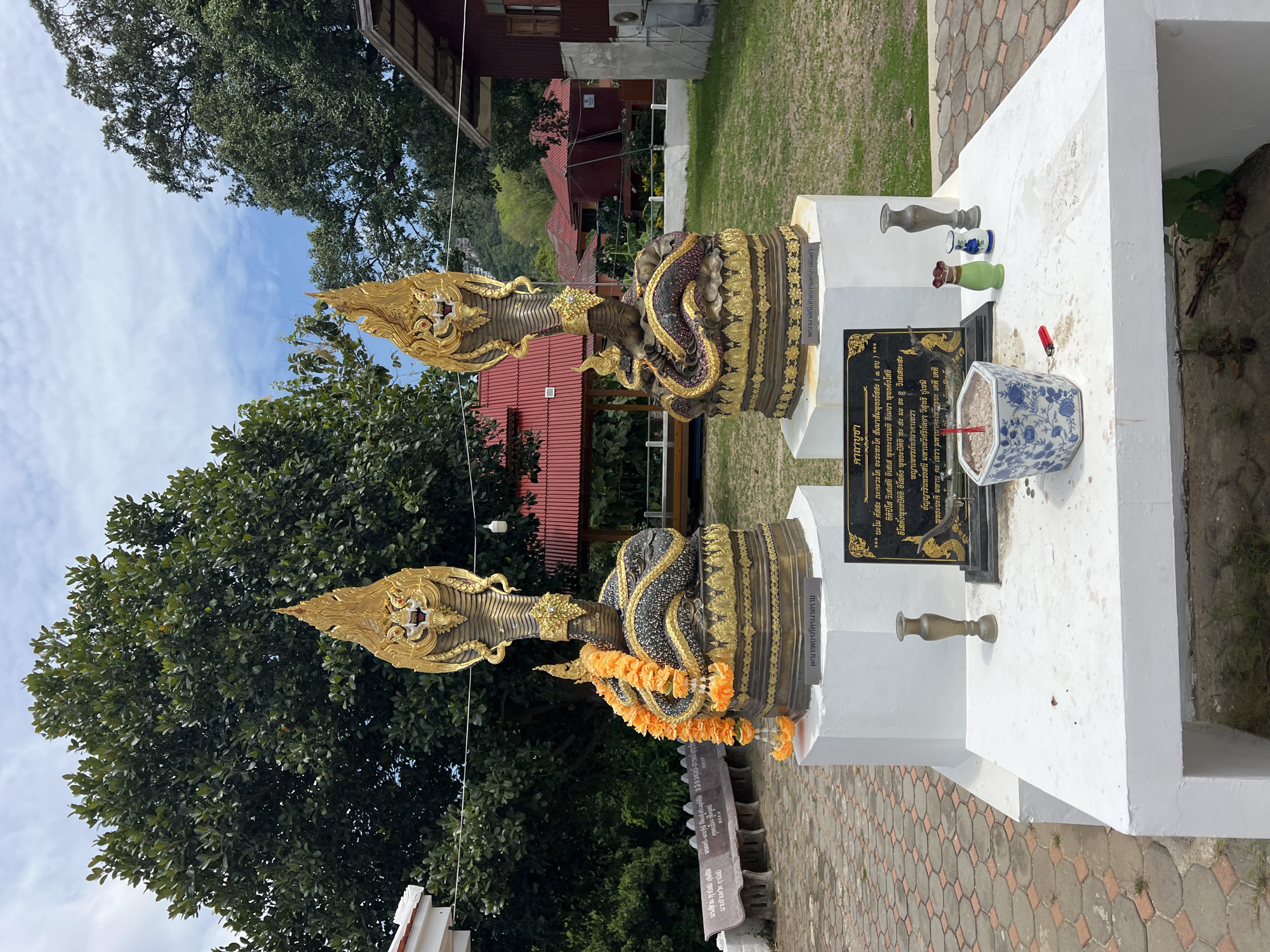 Two Naga statues are situated on a white platform, adorned with yellow flower garlands. A black plaque with golden lettering and a blue and white floral-patterned pot containing incense sticks rest on the platform. Several small vases are also placed nearby. The setting appears to be outdoors, with trees and buildings visible in the background. The location is Baan Hua Thanon, Thailand.