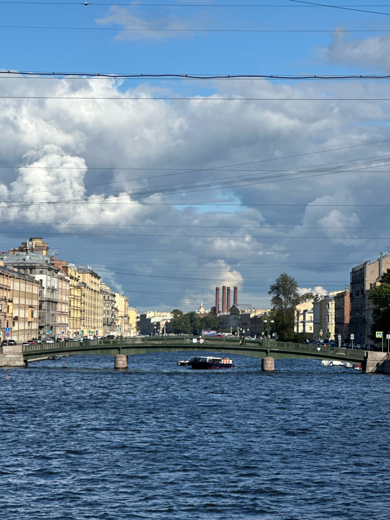 This is a photograph taken from the surface of a body of water, looking towards a city scene. In the foreground, the water is a deep blue with small waves. A green pedestrian and vehicle bridge crosses the water in the mid-ground. The bridge has stone abutments and two visible supports in the water. A narrow, dark-colored boat with a white stripe is moving under the bridge, heading away from the viewer.
Beyond the bridge, on either side of the water, are buildings characteristic of St. Petersburg. The buildings are multi-story structures with varied architectural styles, painted in a range of colors including yellow, beige, and pale pink. On the left bank, the buildings are densely packed, with numerous windows and some visible rooftop details. On the right bank, the buildings are also multi-story, with a more uniform color palette. In the distance, behind the buildings, three tall, red smokestacks are visible. Trees are present between some of the buildings.
The sky above is a mix of bright blue and dramatic, dark grey clouds, suggesting a dynamic weather condition. Several power lines are strung across the upper portion of the image, running horizontally. The overall impression is of a canal or river running through an urban area of St. Petersburg, likely on a day with variable weather.