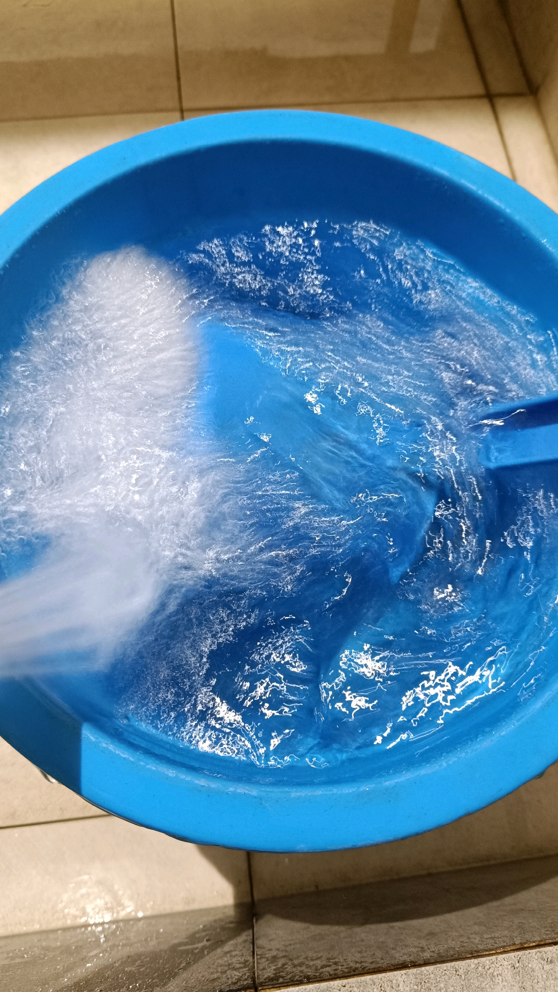 A high-angle shot captures a bright blue plastic basin filled with churning water, likely being filled from a tap. The water appears frothy and turbulent, suggesting a strong flow. The basin is resting on a tiled floor, with grout lines visible. The tiles are a neutral, light brown color. A small portion of a blue plastic object, possibly a scoop or handle, is submerged in the water on the right side of the frame. The lighting suggests an indoor setting, possibly a bathroom or laundry area. There are no people or animals visible in the image. The scene is devoid of any specific landmarks that would indicate the city of Tomohon, Indonesia.
