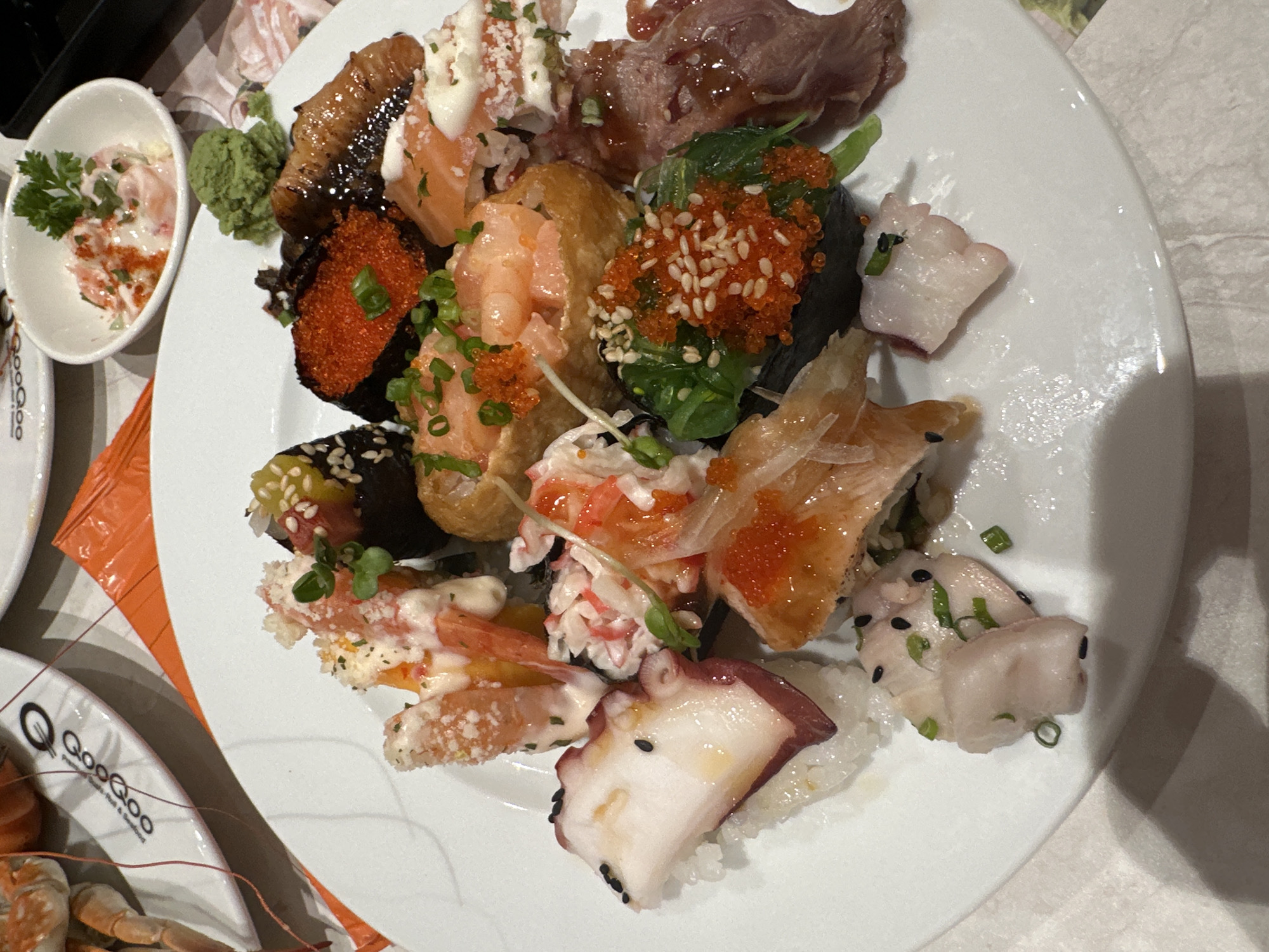 A high-angle shot shows a white plate densely packed with various types of sushi. The assortment includes nigiri topped with octopus, salmon with mayonnaise, eel, and a type of meat. There is also sushi wrapped with seaweed and garnished with tobiko, shrimp with tobiko, and inari sushi topped with shrimp. The sushi is decorated with scallions and sprouts. Beside the main plate, a small bowl contains what appears to be seafood salad. Wasabi is situated near the side of the plate. Additional plates, one of which is branded "QooQoo," and a serving of shellfish are partially visible in the lower left of the frame. A patterned placemat is visible under the plates, and a textured surface, likely a tablecloth, forms the background. The location is identified as Nha Trang Ward, Vietnam.