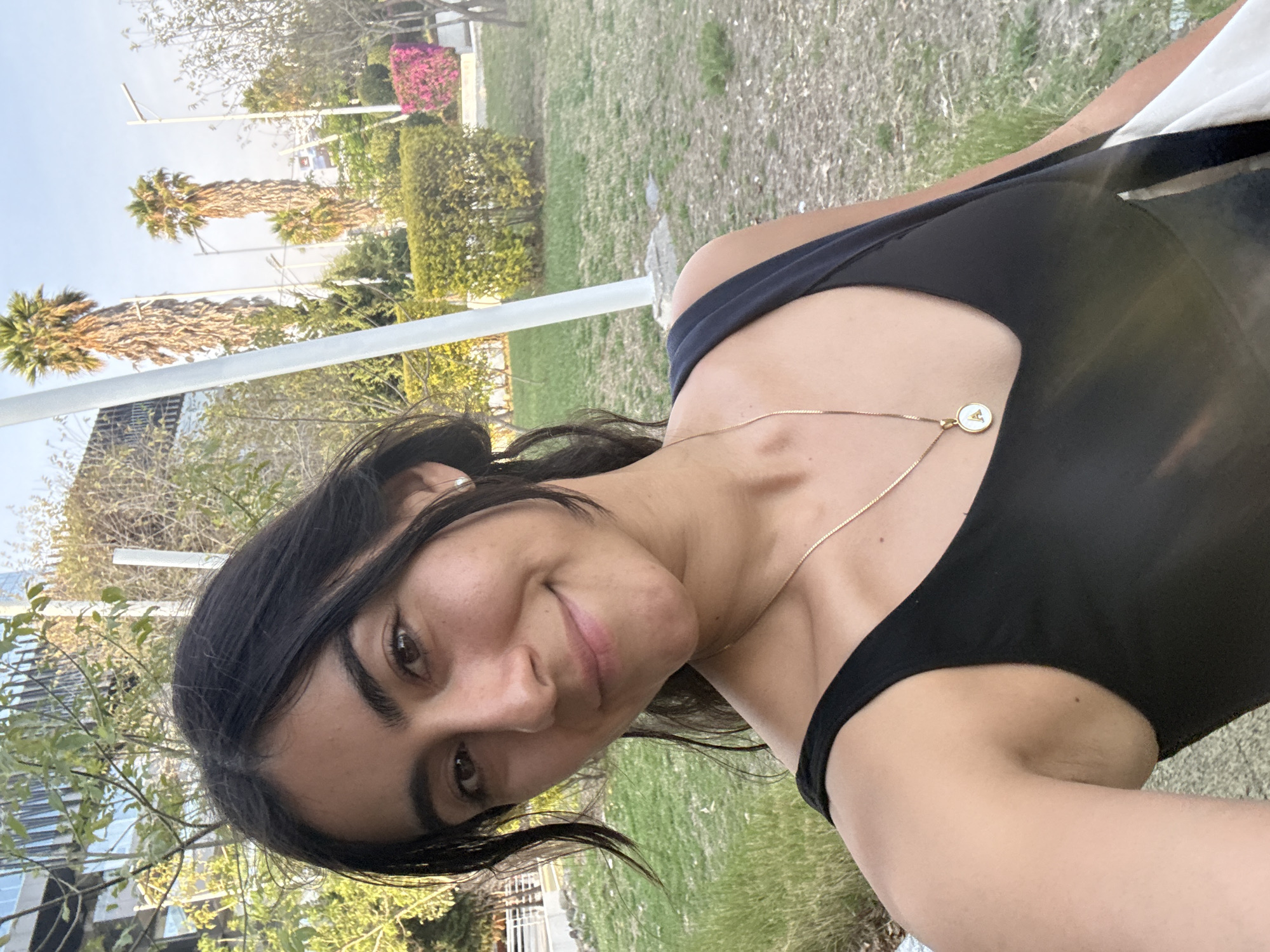 A woman with dark hair and brown eyes is positioned in the foreground. She is wearing a black top and a gold necklace with a circular pendant. The background features a park-like setting with green grass, trees, and palm trees. In the distance, there are buildings and a clear sky. The scene is in San Bernardino Tlaxcalancingo, Mexico.
