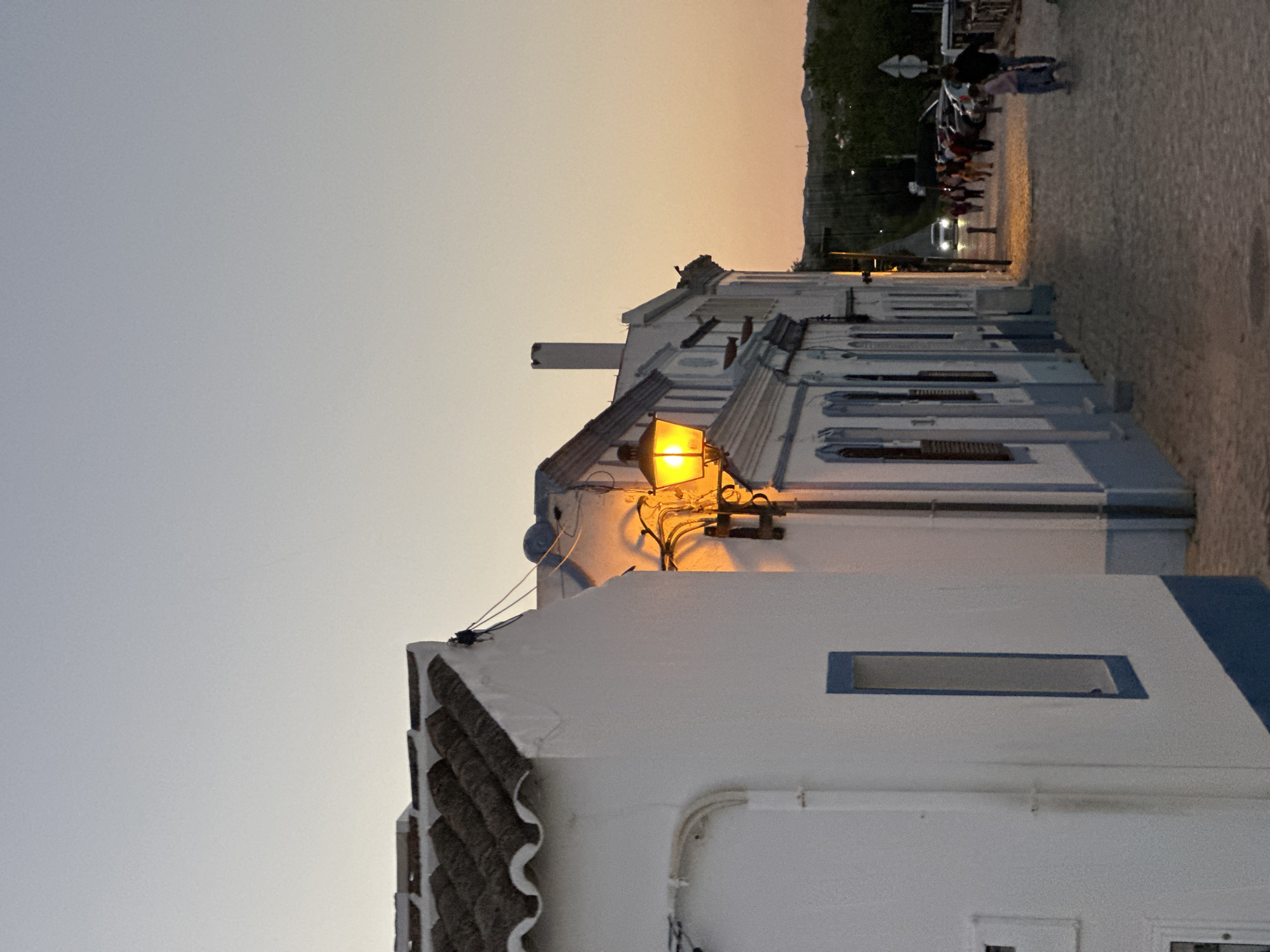 Captured from a ground-level perspective, the image showcases a street in Vila Real de Santo António, Portugal, during twilight. A row of white buildings with blue accents extends into the depth of the image, running parallel to a pedestrian walkway. A bright orange light fixture affixed to one of the buildings illuminates the immediate surroundings. A group of pedestrians and parked vehicles are visible further down the street. The sky transitions from a gradient of orange near the horizon to a pale grey closer to the top of the frame. The buildings are predominantly white with tiled roofs.