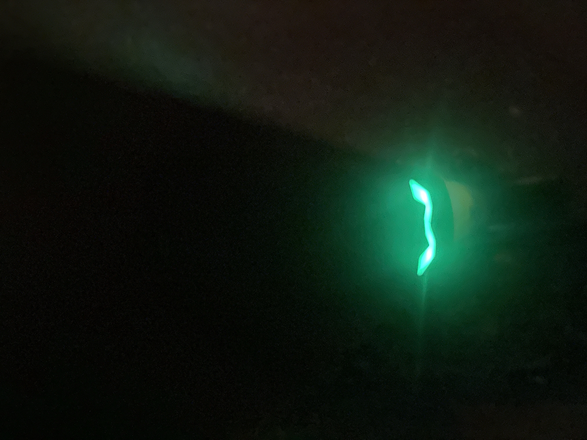 The image is a close-up, low-light photograph of a green light source against a dark background. The light has a distinct, curved shape, resembling a smile or a stylized eyebrow. There are no discernible people, objects, or environmental features other than the light and the dark background. The location in the city of Minsk, Belarus, cannot be determined from the image.
