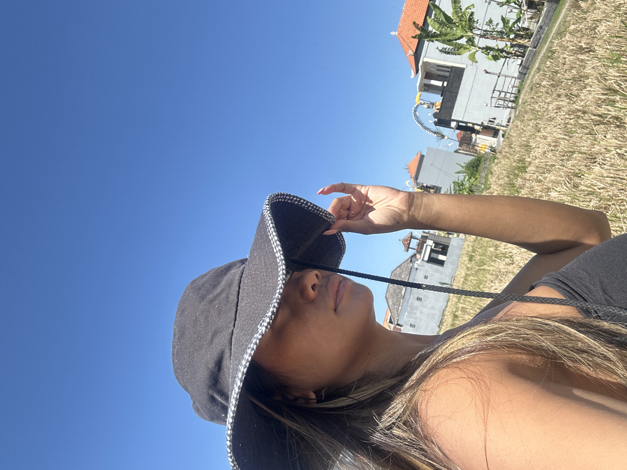 A person wearing a black bucket hat with a checkered band is lying down in a field of dry grass under a clear blue sky. The hat casts a shadow over their face, obscuring most of their features, but their chin and lips are visible. Their right hand, with bright pink fingernails, is holding the brim of the hat. Long, blonde-streaked hair fans out around their shoulder and neck.
In the background, several buildings with red-tiled roofs are visible. Some of these appear to be houses or small structures, with one larger building to the right featuring multiple levels and balconies. Banana trees with large green leaves grow near one of the buildings. Decorative elements, possibly religious or festive, are also present, including a curved archway adorned with white circular ornaments and what looks like flags or streamers. The dry grass suggests a time of year when crops have been harvested or the land is arid.
The bright sunlight and clear sky indicate it is daytime, likely midday or afternoon. The overall impression is one of relaxation and enjoying the outdoors in a tropical or sub-tropical location, possibly Sukawati, Indonesia, given the architectural style and vegetation. There is no visible text in the image.