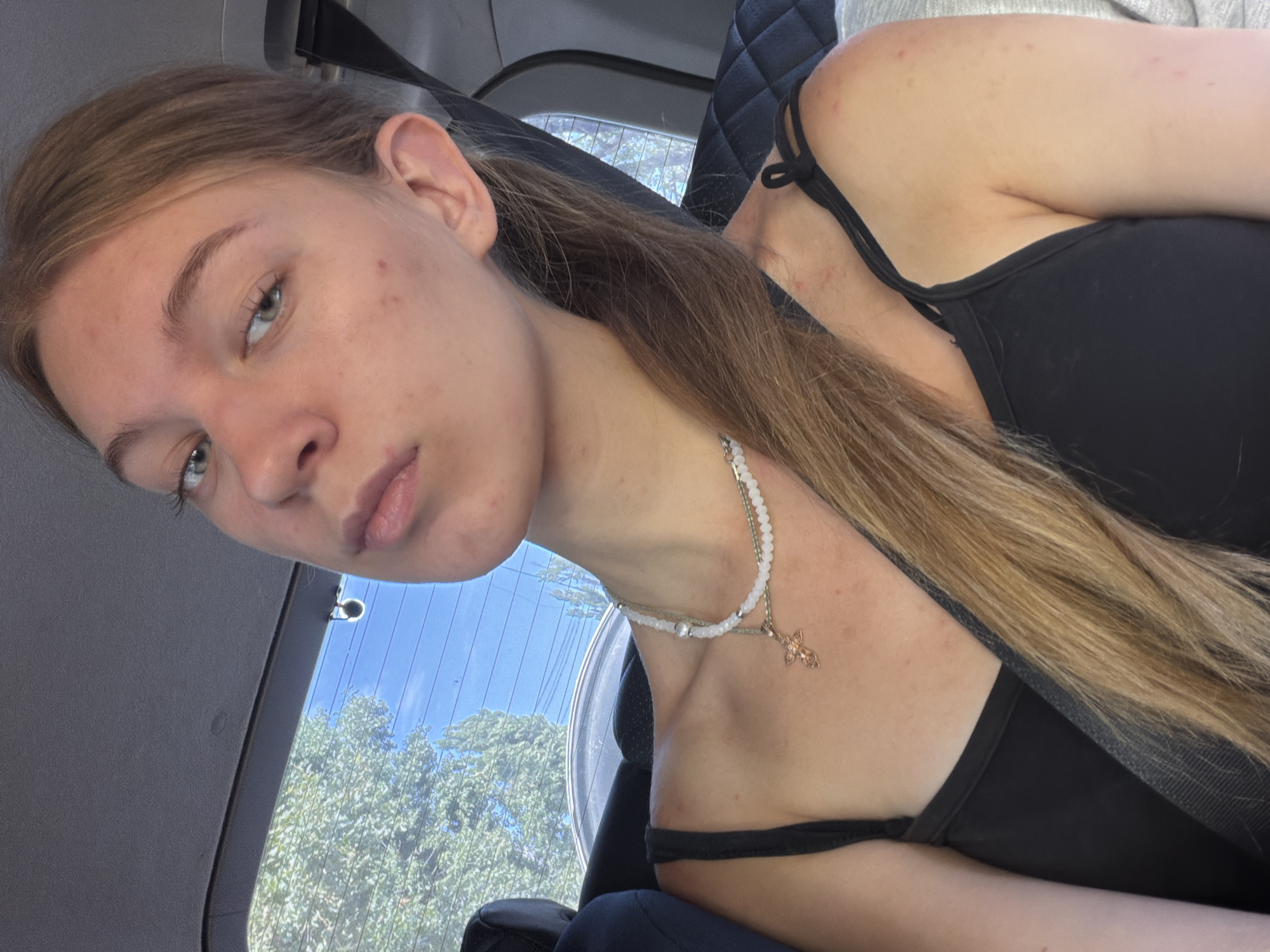A young Caucasian woman with light brown hair pulled back into a ponytail is taking a selfie inside a vehicle. She is wearing a black tank top and a beaded necklace with a small cross pendant. The background shows a car's interior with a seat and a rear window revealing trees and a blue sky. The image appears to be taken outdoors during the daytime. There is no discernible information in the image to indicate a specific location in Rostov-on-Don, Russia.