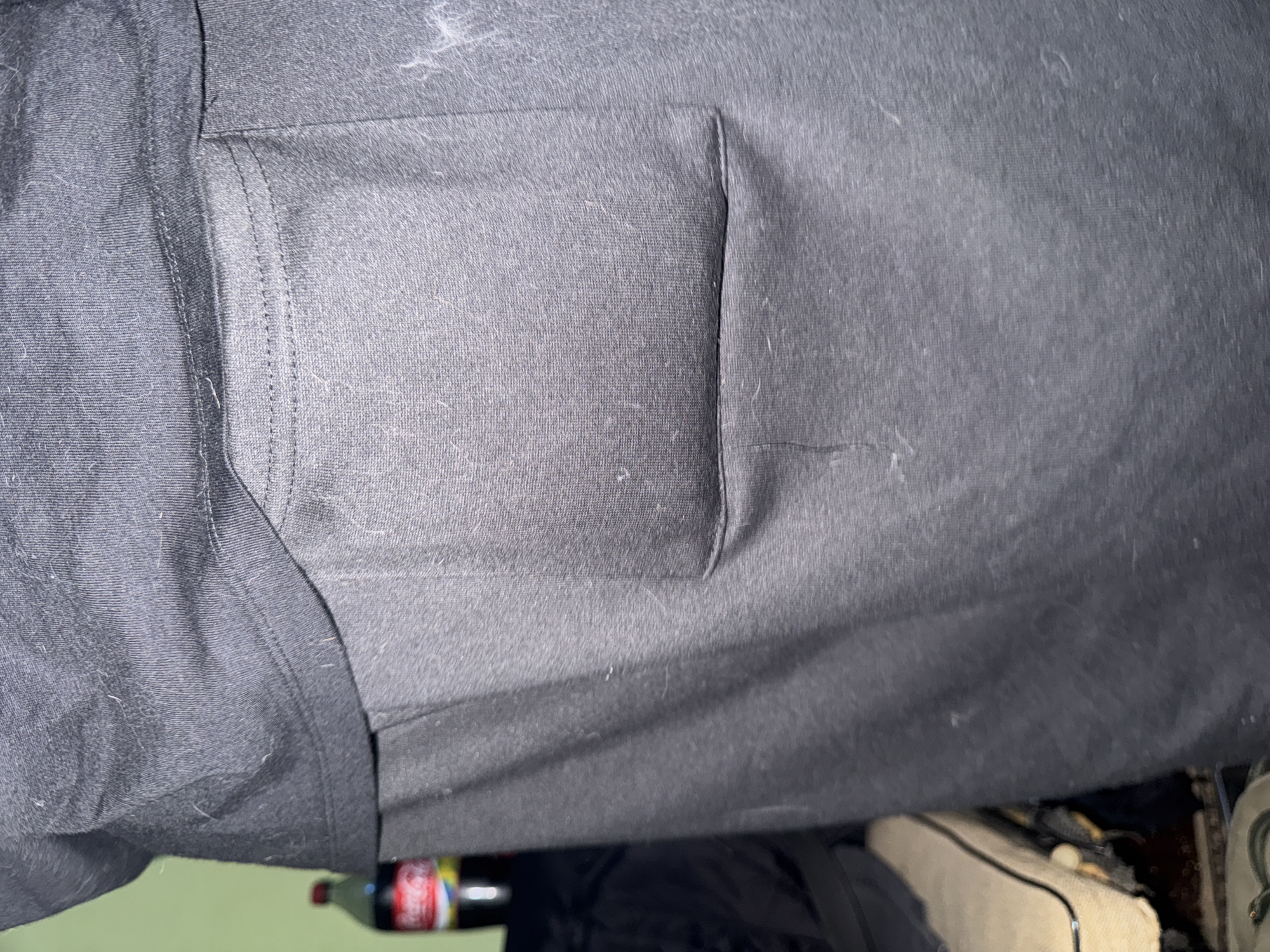 A close-up image shows a black shirt with a pocket on the left breast. The fabric has visible fibers and some lighter-colored lint. The lower part of the frame reveals the tops of a Coca-Cola bottle and a light-colored bag. The background includes a green surface and other indiscernible items.