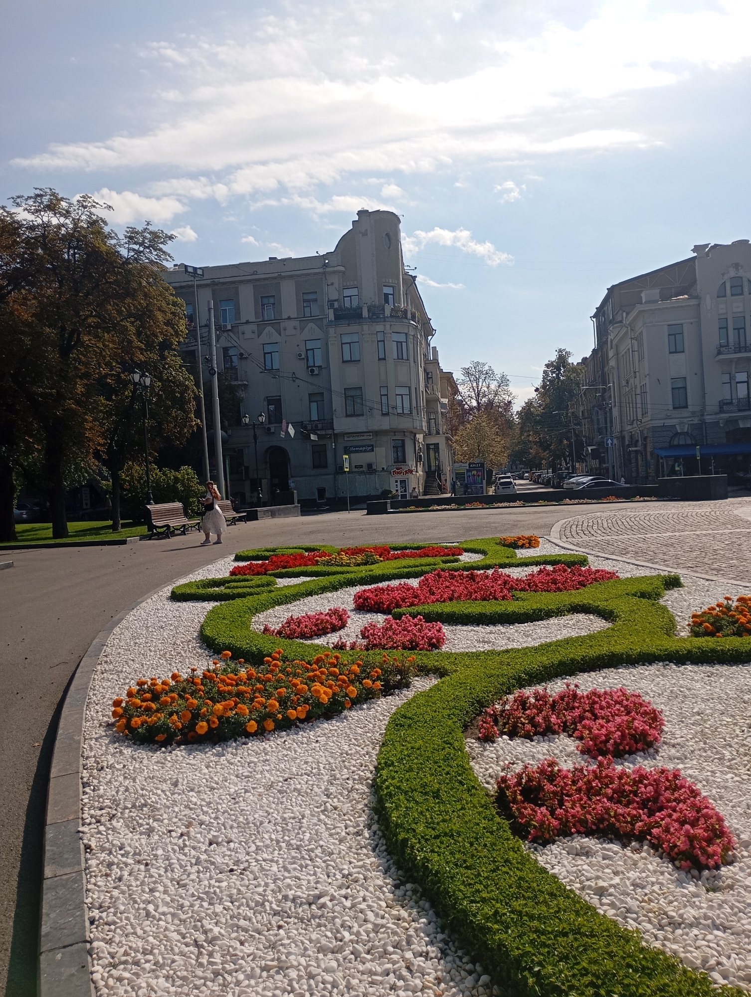 This is a ground-level, exterior, daytime photograph taken in Kharkiv, Ukraine. The foreground features a large, ornamental flowerbed with a serpentine design. The flowerbed is filled with white pebbles, low-lying green bushes, and patches of vibrant pink and orange flowers. A woman in a light-colored dress is walking in the middle ground, to the left of the flowerbed, towards the left edge of the frame. Behind her, there are two park benches situated in front of a row of mature trees with autumnal foliage. Beyond the trees and benches, an urban street scene unfolds with several multi-story buildings in various architectural styles. Cars are visible parked along the street and in motion. The sky is bright blue with scattered white clouds, indicating clear weather.