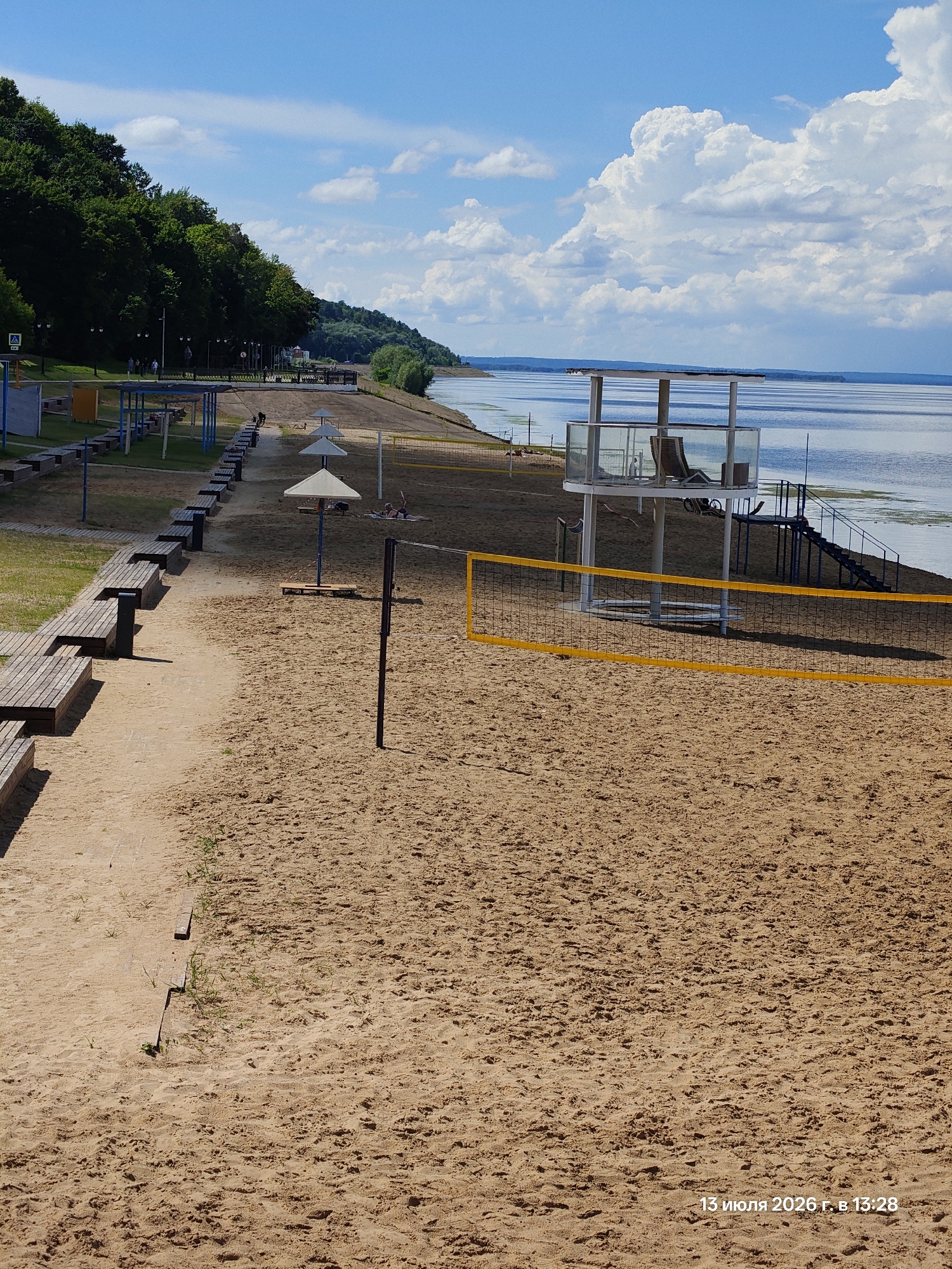 The image depicts a sandy beach with a volleyball net and several beach umbrellas. In the background, there is a body of water and a dense green treeline. A structure with a glass railing and a chair is visible on a raised platform. A date and time stamp in Russian, "13 июля 2026 г. в 13:28", is present in the bottom right corner. The scene appears to be a public recreational area.