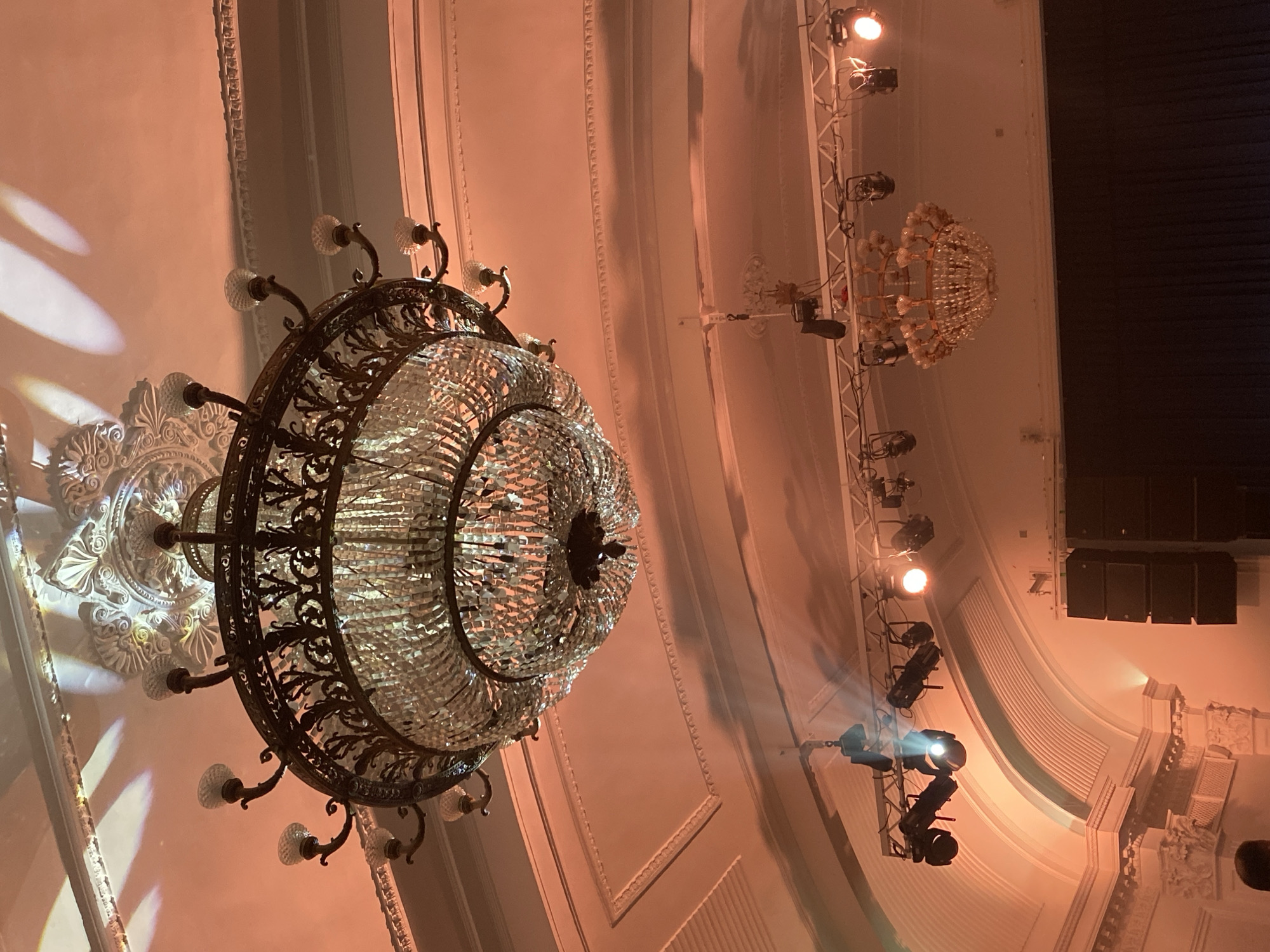 A low-angle shot depicts the interior of what appears to be a theater or grand hall, likely in Kazan, Russia, based on architectural styles common in the region. Two ornate chandeliers dominate the ceiling, the larger one closer to the viewer with a complex ironwork frame and numerous glass or crystal elements. The smaller chandelier is visible further back and to the right. Stage lighting fixtures mounted on vertical trusses are also present, one of which is actively projecting a beam of light. The walls have decorative molding and are lit with a warm, orange hue. A speaker array is visible in the upper right, near a dark, possibly curtained area.