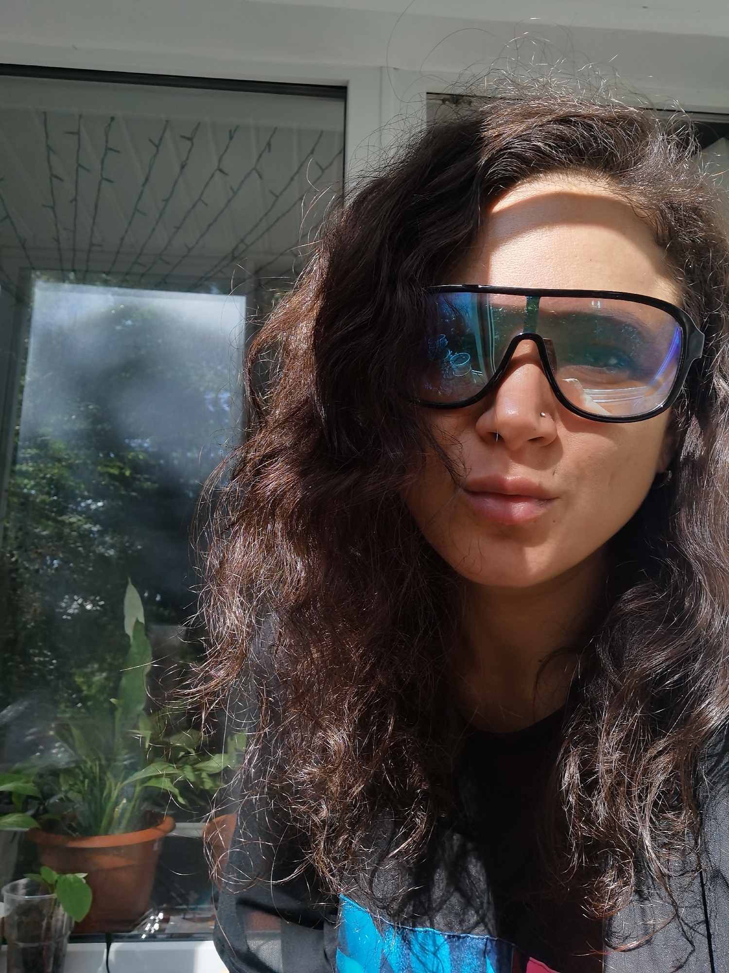 A woman with dark, curly hair and glasses is looking at the camera. She is wearing a black shirt. The background shows a window with plants and string lights. The scene is lit by sunlight. The location is in the city of Chaikovsky, Russia.