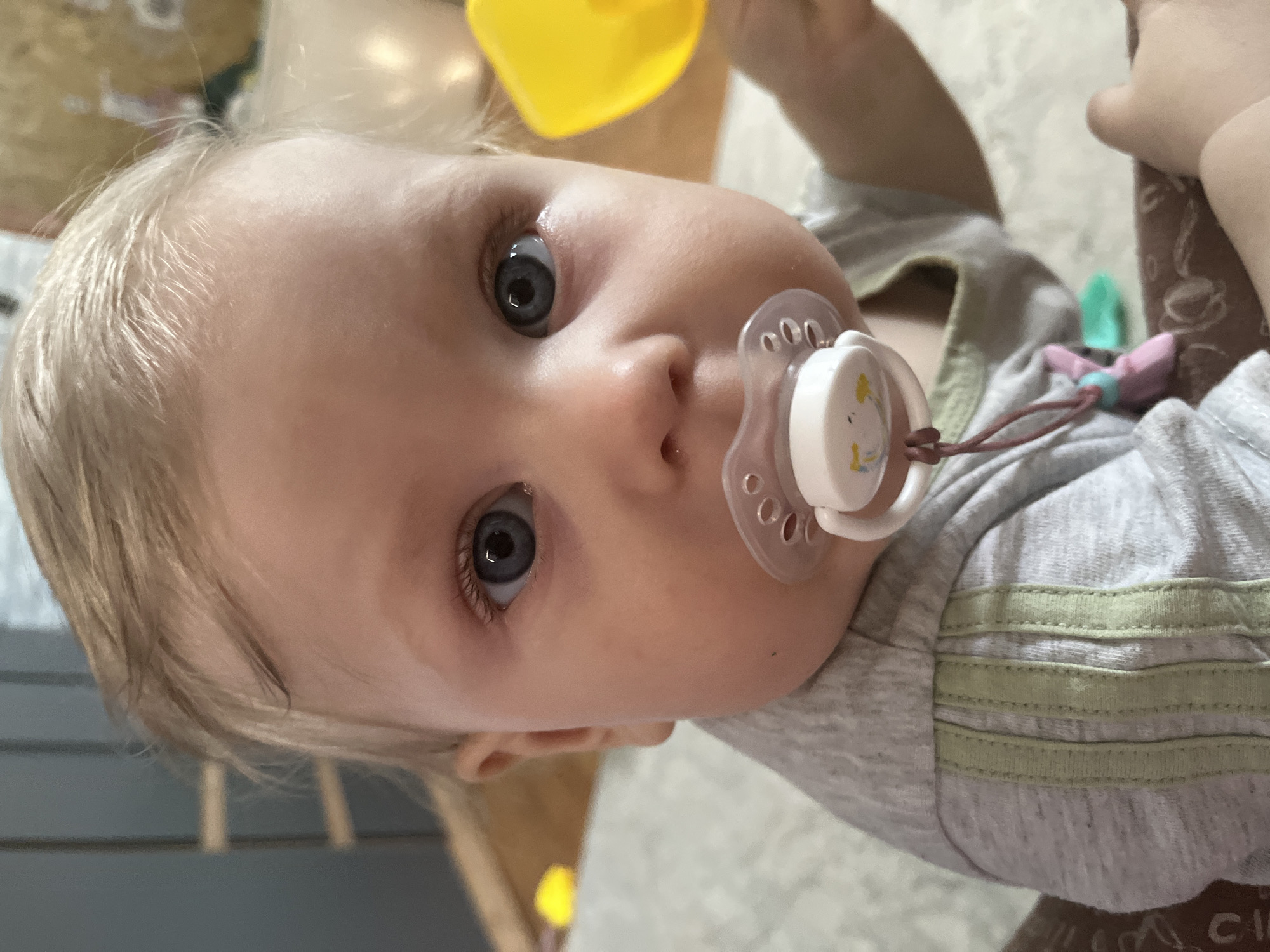 A close-up, high-angle shot shows a baby with fair skin, light blonde hair, and wide, deep blue eyes, looking directly into the camera. The baby is wearing a grey t-shirt with three light green horizontal stripes on the sleeve, visible in one of the crops. A white pacifier, featuring a small cartoon animal design (possibly a duck or bunny) on its button, is in the baby's mouth. The pacifier is connected to a string or clip, which has pink and blue elements, extending towards the baby's chest.
In the background, blurred elements suggest an indoor setting. A yellow object, possibly a toy or bottle, is partially visible near the top of the baby's head, held by an adult hand. The baby's expression is neutral and observant. The full image is rotated, implying the baby is lying down. The scene depicts a quiet moment, likely at home in Vesyoloye, Russia, with the baby either resting or engaging with an adult. Faint, partial, illegible text, possibly "CI", can be seen on a dark, patterned object in the bottom right corner of one of the crops.