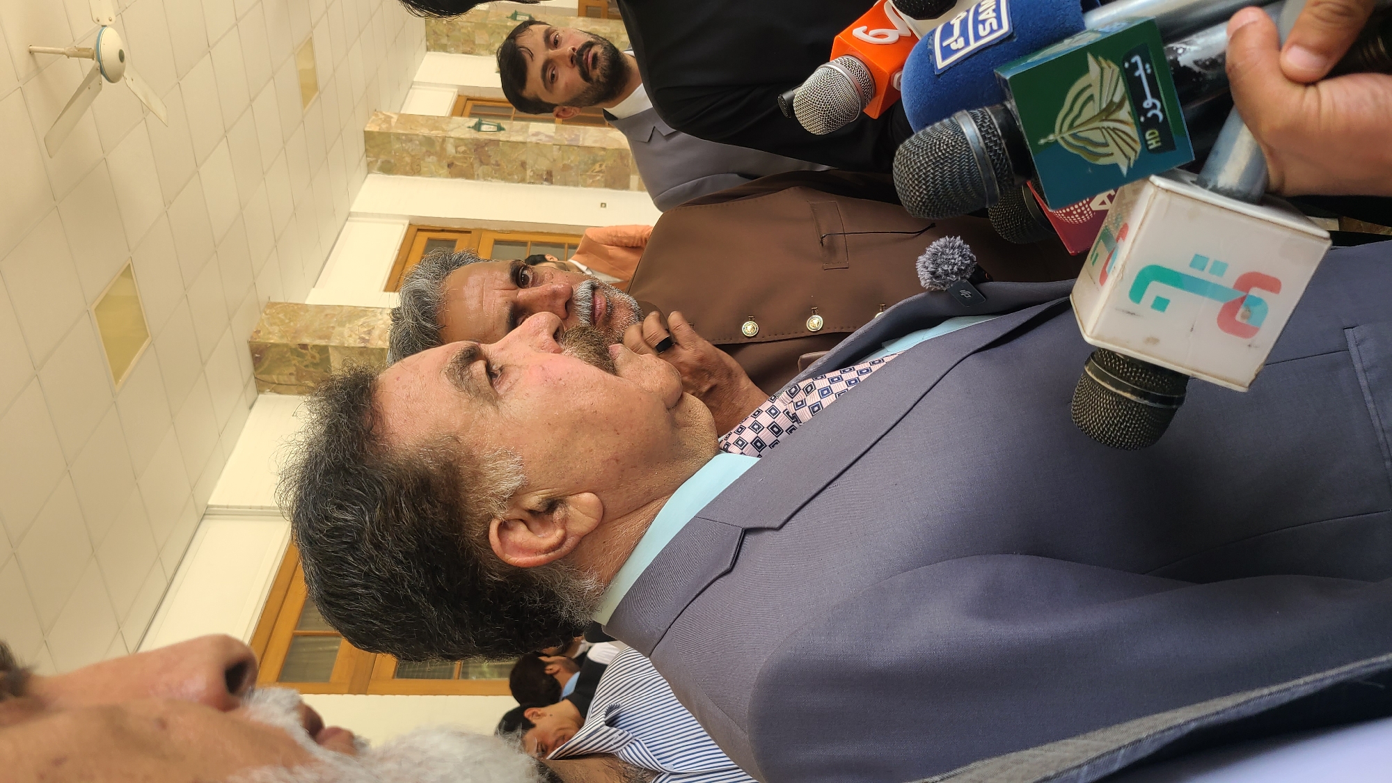 The image appears to depict a press conference or media event, possibly occurring outdoors, given the natural light. Several men are present. A man in a grey suit appears to be the focal point, as microphones from various news outlets are positioned close to him. Another man in a brown waistcoat stands behind him. There is a third man with a black suit in the back. The location is described as Quetta City Tehsil, Pakistan.