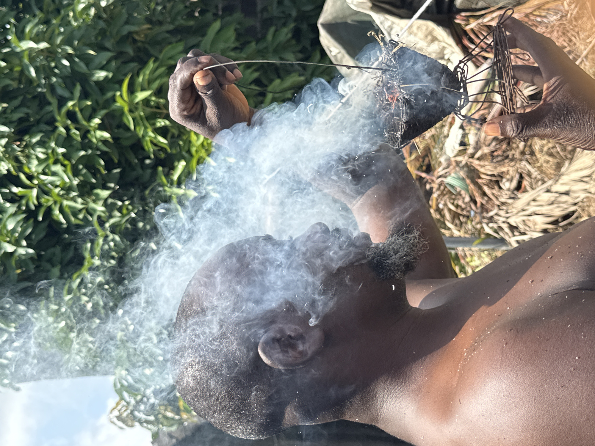 A close-up shot focuses on a shirtless Black man, his head tilted back, enveloped in a cloud of grey smoke. He holds a smoking metal object near his mouth with his right hand and a similar object with his left, held higher in the frame. The background includes green foliage and dried plant matter. The location appears to be outdoors, possibly in Ado, Nigeria, based on the request. The man's skin is glistening, suggesting possible moisture. The scene centers around the act of inhaling smoke from a burning object, with the surrounding elements contributing to the overall atmosphere.