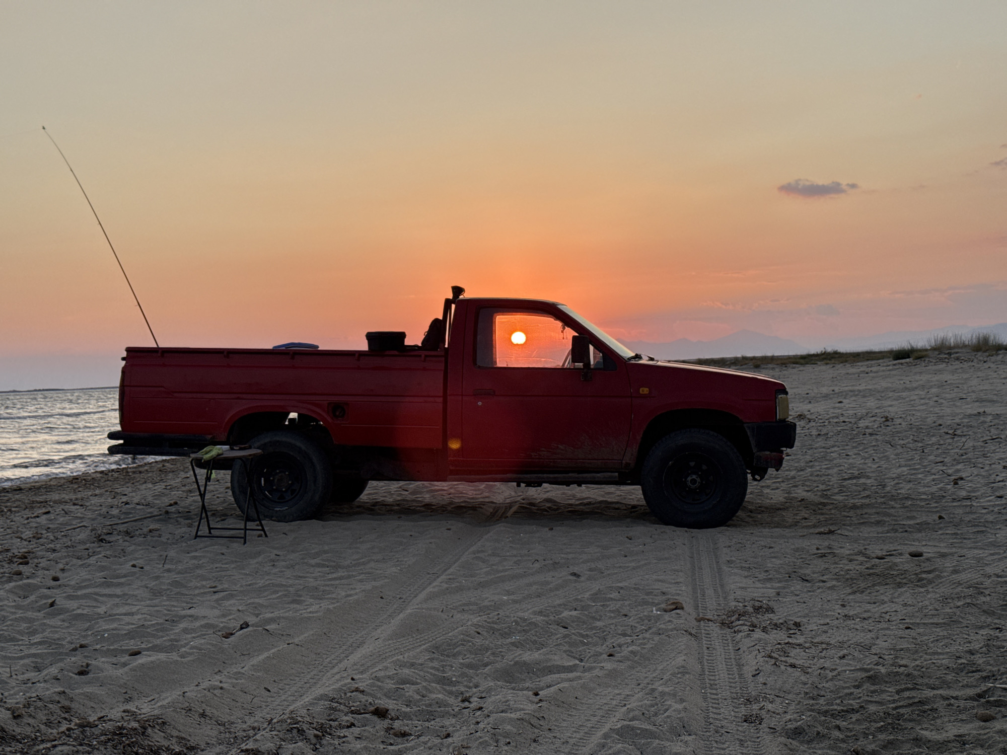 A red pickup truck is parked on a sandy beach in Imeros, Greece, at sunset. The sun is visible through the driver's side window, casting a warm glow. A fishing rod is propped up behind the truck, and a small folding stool is placed near the rear tire. The ocean waves are gently lapping at the shore in the background, and distant mountains are silhouetted against the orange and yellow sky. Tire tracks are imprinted in the sand, indicating the truck's presence. The overall scene depicts a peaceful, serene moment at the end of the day.