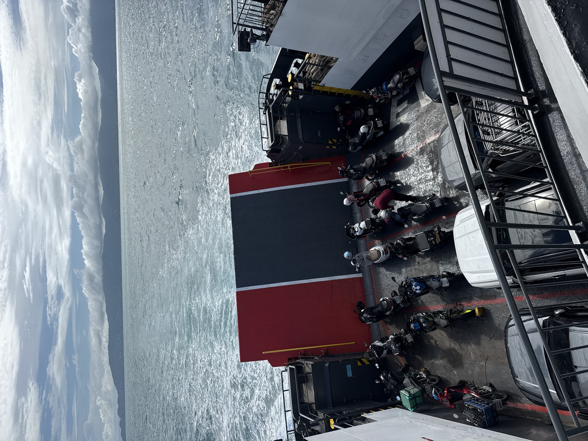On a ferry in the waters near Unknown, Thailand, the open deck is filled with parked motorcycles and two cars. The motorcycles are varied in color and type, some with storage boxes or riders, while the cars are dark in color. The ferry is moving, indicated by the wake in the water and the choppy sea. In the background, a mountainous coastline under a cloudy sky is visible. The deck itself is wet, possibly from rain or sea spray, with red and black painted lines demarcating parking spots. A large red and black gate or ramp is positioned on the deck.
