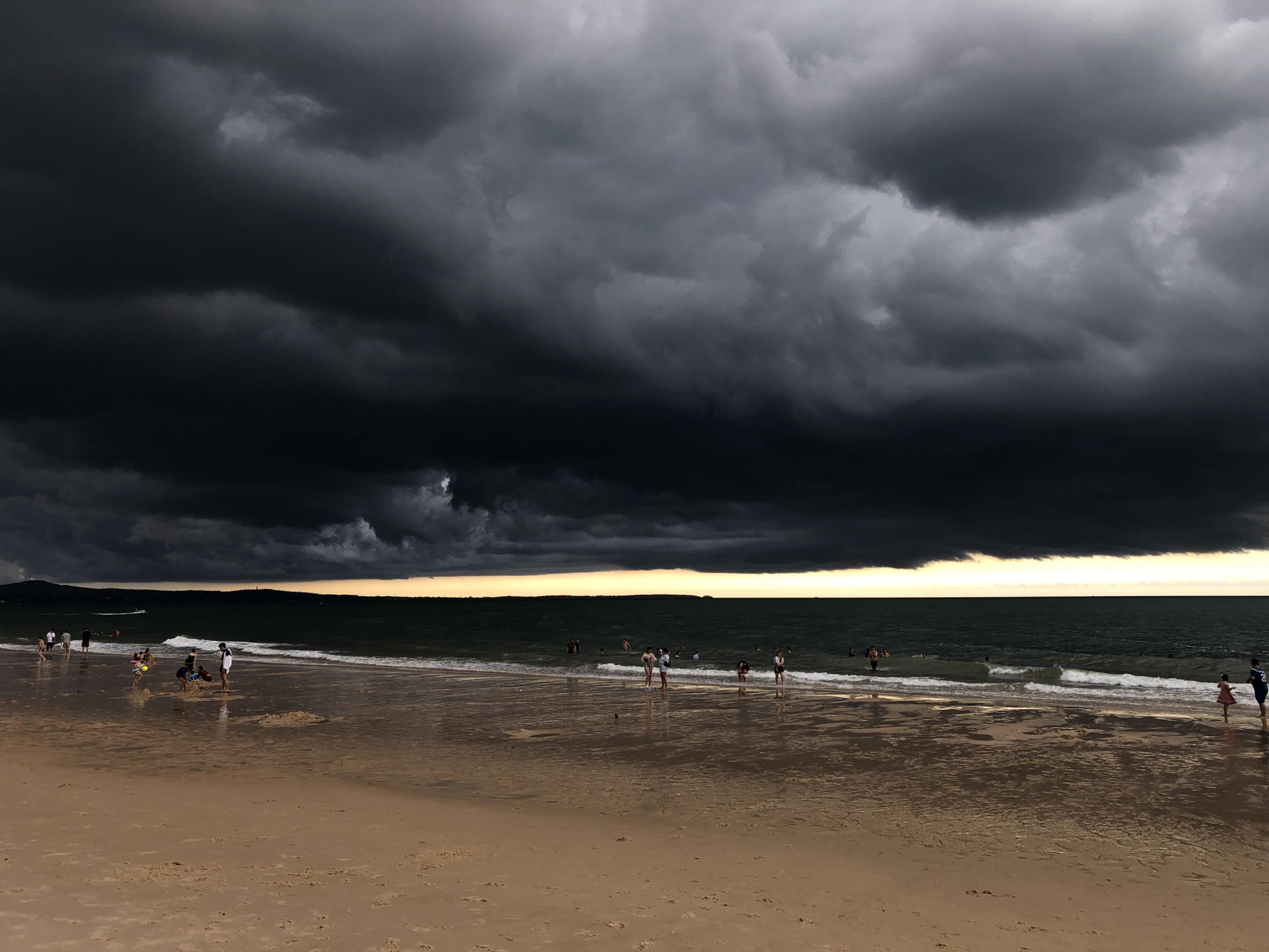 The image depicts a beach scene in Phu Thuy Ward, Vietnam, under a dramatic and ominous sky. A vast expanse of dark, heavy storm clouds dominates the upper portion of the frame, casting a somber mood over the scene. The clouds appear dense and textured, with a gradient from deep black at the top to lighter shades of gray closer to the horizon.
Below the clouds, the sea is a dark, choppy green, with white waves breaking near the shore. A thin band of pale yellow light is visible along the horizon, separating the sea from the clouds, suggesting either the very late stages of sunset or the early approach of a storm.
On the sandy beach in the foreground, several people are visible. Some are wading in the shallow water, while others are standing on the wet sand closer to the sea. Further up the beach, near the dry sand, a few individuals are scattered, some appearing to be children playing. The wet sand reflects the muted light, creating shimmering patterns. A small boat is visible in the distance on the water, and a strip of land with hills can be seen along the distant coastline. The overall impression is one of a beach occupied by a few individuals as a significant weather event approaches.