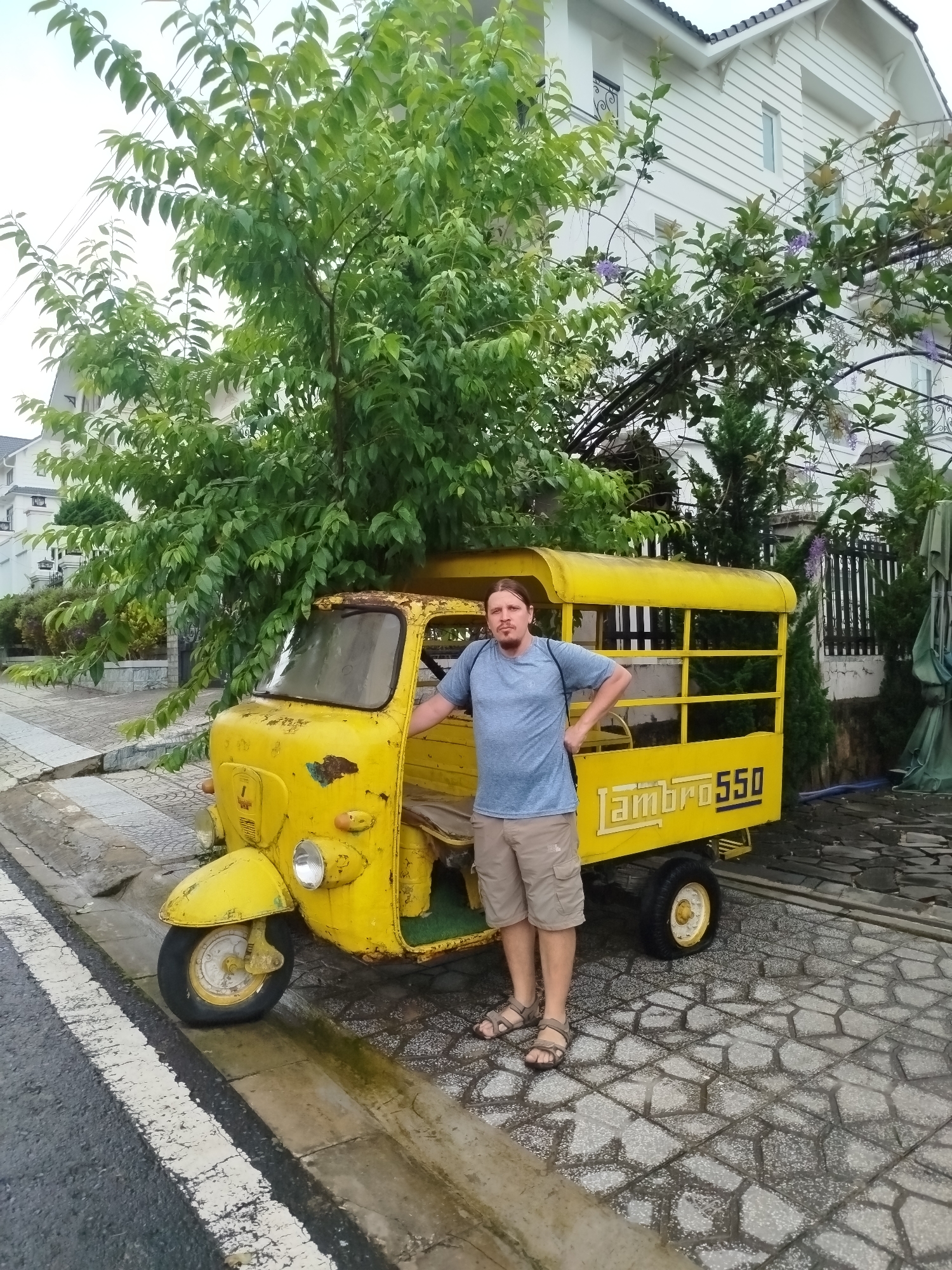 A man stands next to a yellow, three-wheeled vehicle, possibly a Lambro 550, which is parked on a stone-paved walkway. The man, wearing a blue t-shirt and khaki shorts, has his hands on his hips. He is positioned on the right side of the vehicle, facing the camera. The vehicle has a weathered appearance, with rust visible on its front. Behind the vehicle, a white building with a dark roof and windows is visible. Lush green trees and foliage surround the scene, with some purple flowers on an overhead structure. The walkway is wet, suggesting recent rain. This scene appears to be in the city of Da Lat, Vietnam, known for its French colonial architecture and temperate climate.