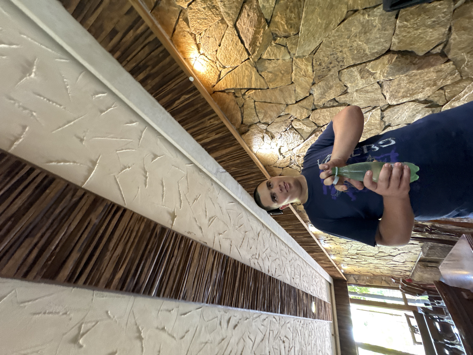 A young man is standing in a room with a stone wall and a wooden ceiling, holding a green bottle of water. The ceiling is decorated with wooden beams and white textured panels. The wall is made of large, irregularly shaped stones, with warm lighting illuminating it. To the left, there are tables and chairs, suggesting the room is a restaurant or dining area. Natural light streams in from a window on the far left. The man is wearing a dark blue t-shirt with a graphic design and appears to be in the process of opening the bottle. The scene is set in Sredets, Bulgaria.
