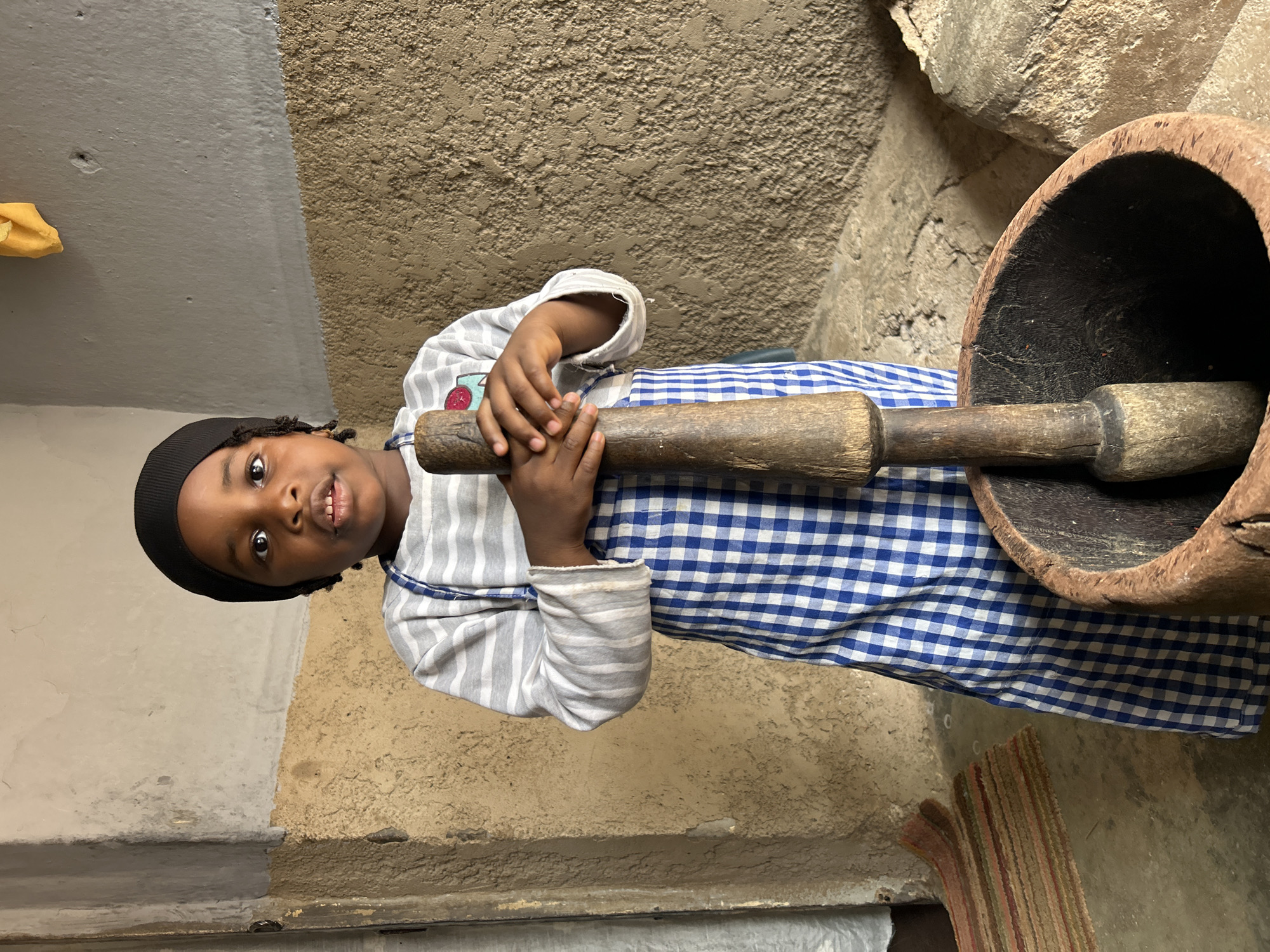 A young child is standing in what appears to be a room in Jos, Nigeria. The child is holding a large wooden pestle that is situated within a wooden mortar. The child is wearing a black cap, a striped, long-sleeved shirt, and a blue and white checkered dress or apron. The background consists of walls with varying textures and colors, suggesting an older structure. A rug is visible on the floor. The mortar appears to be a traditional African grinding tool.