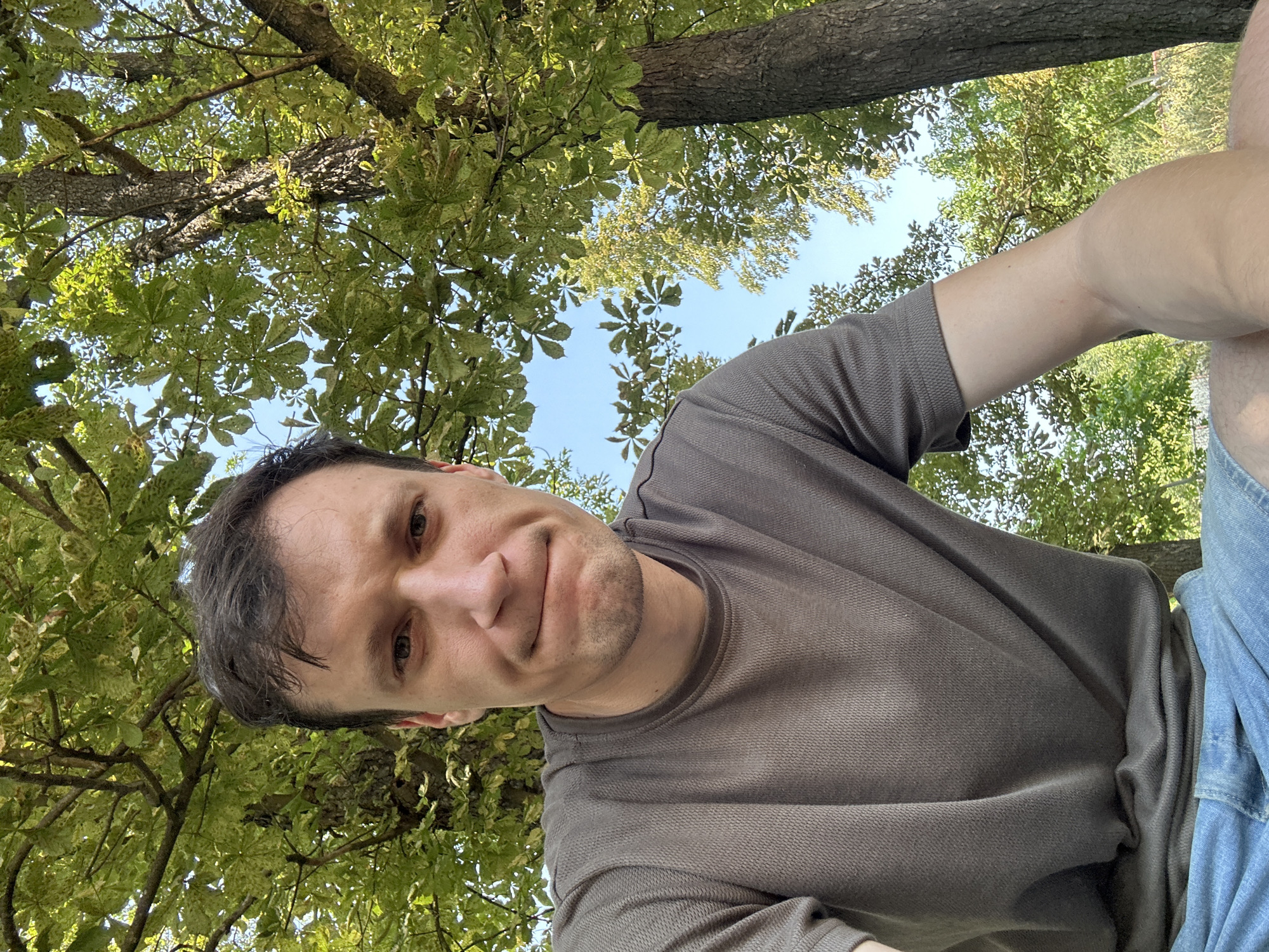 A close-up selfie of a man with short brown hair and blue eyes, wearing a gray t-shirt and blue denim shorts. He is looking directly at the camera with a slightly furrowed brow and a subtle smirk. The background features lush green trees with dappled sunlight filtering through the leaves, suggesting an outdoor setting. The sky is visible through the branches, appearing clear and blue. The image was taken in Makiv, Ukraine.