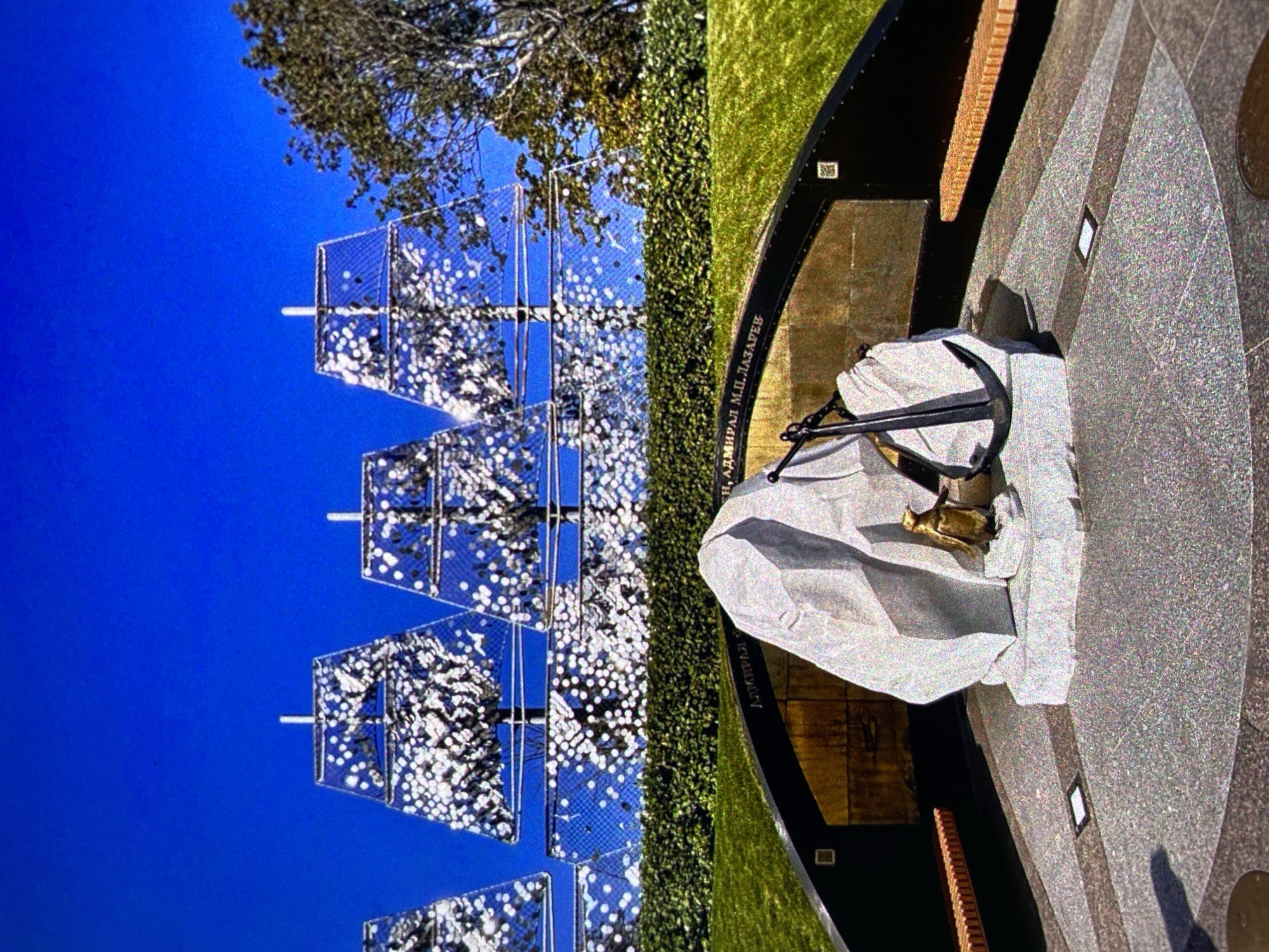 The image shows a monument dedicated to Admiral Mikhail Lazarev in Saint Petersburg, Russia. The monument features a granite base with a covered bust of the admiral and a dark metal anchor resting on top. Surrounding the base is a semicircular structure with a low, green wall and plaques inscribed with Russian text, including the name "Адмирал М.П. Лазарев" (Admiral M.P. Lazarev). In the background, there is an artistic structure with what appear to be mirrored or reflective panels, set against a clear blue sky. A tree is visible partially obscuring the mirrored structure.