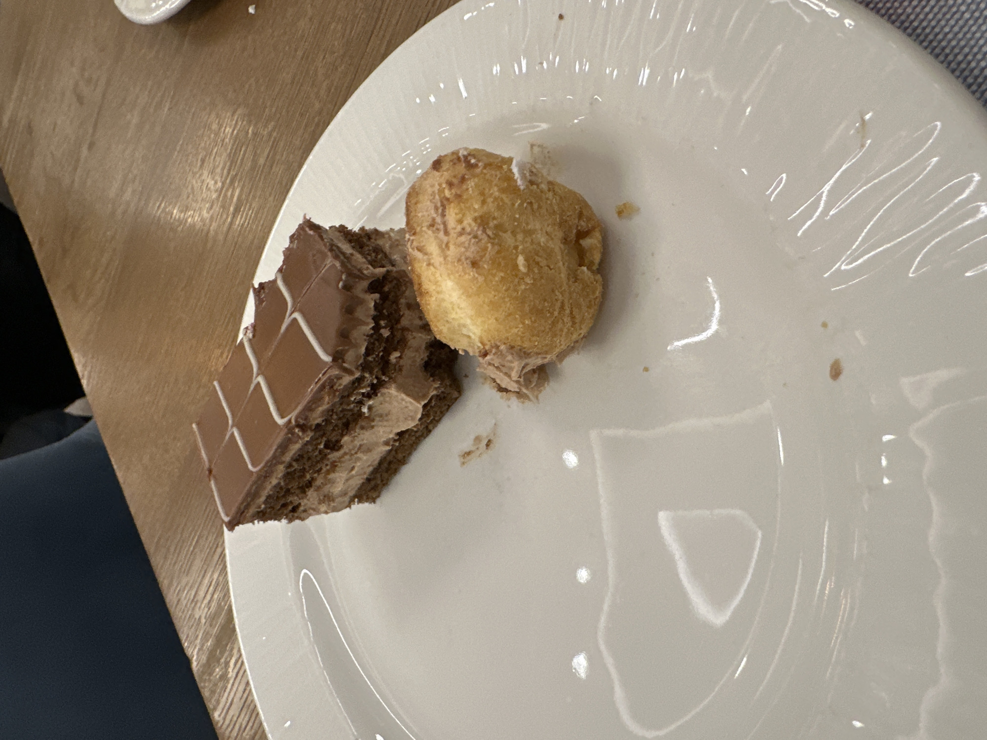 A slice of chocolate cake and a cream puff are on a white plate on a wooden table. The cake has a brown chocolate topping with white icing in a grid pattern, and a layer of cream filling. The cream puff is round and golden brown. The location is unknown and cannot be determined from this image.